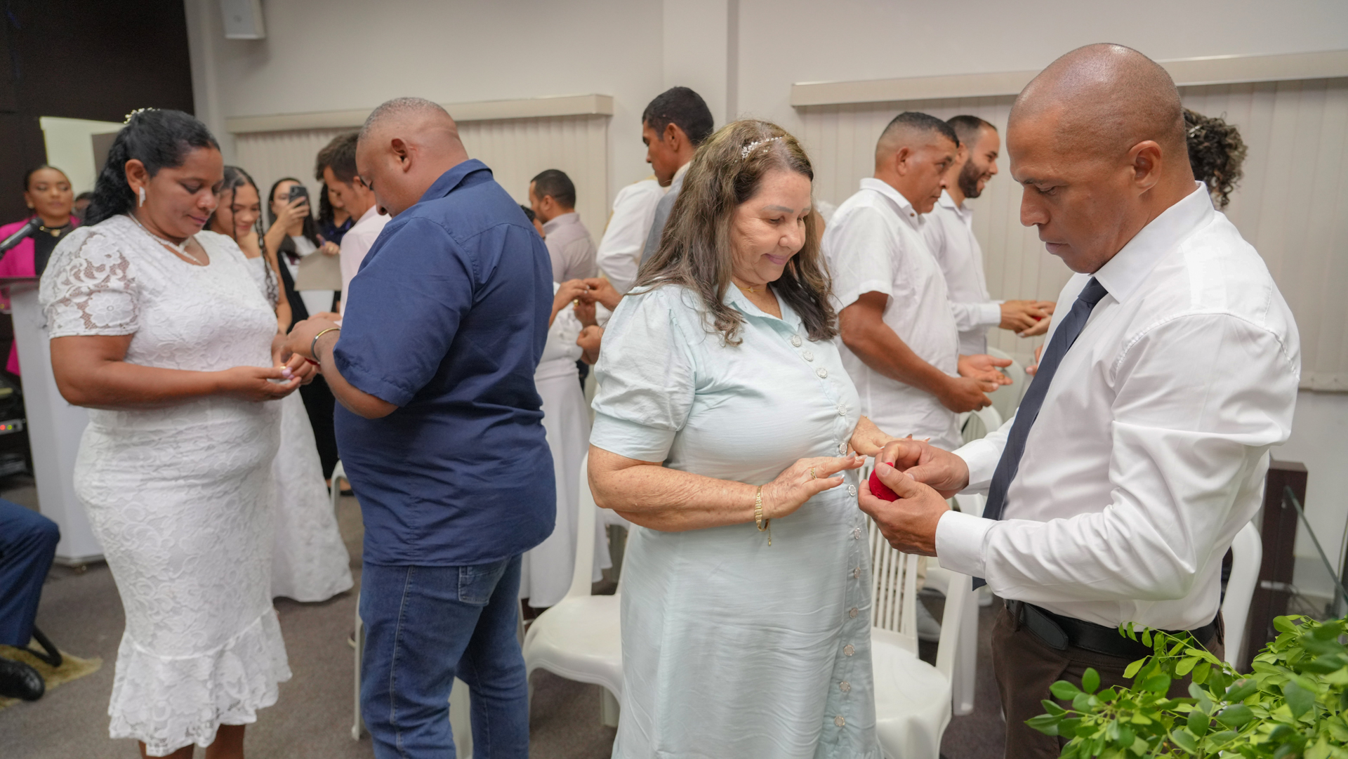 Momento simbólico durante cerimônia coletiva: casais participam de troca de alianças, em ambiente fechado e organizado, acompanhados por familiares e convidados.