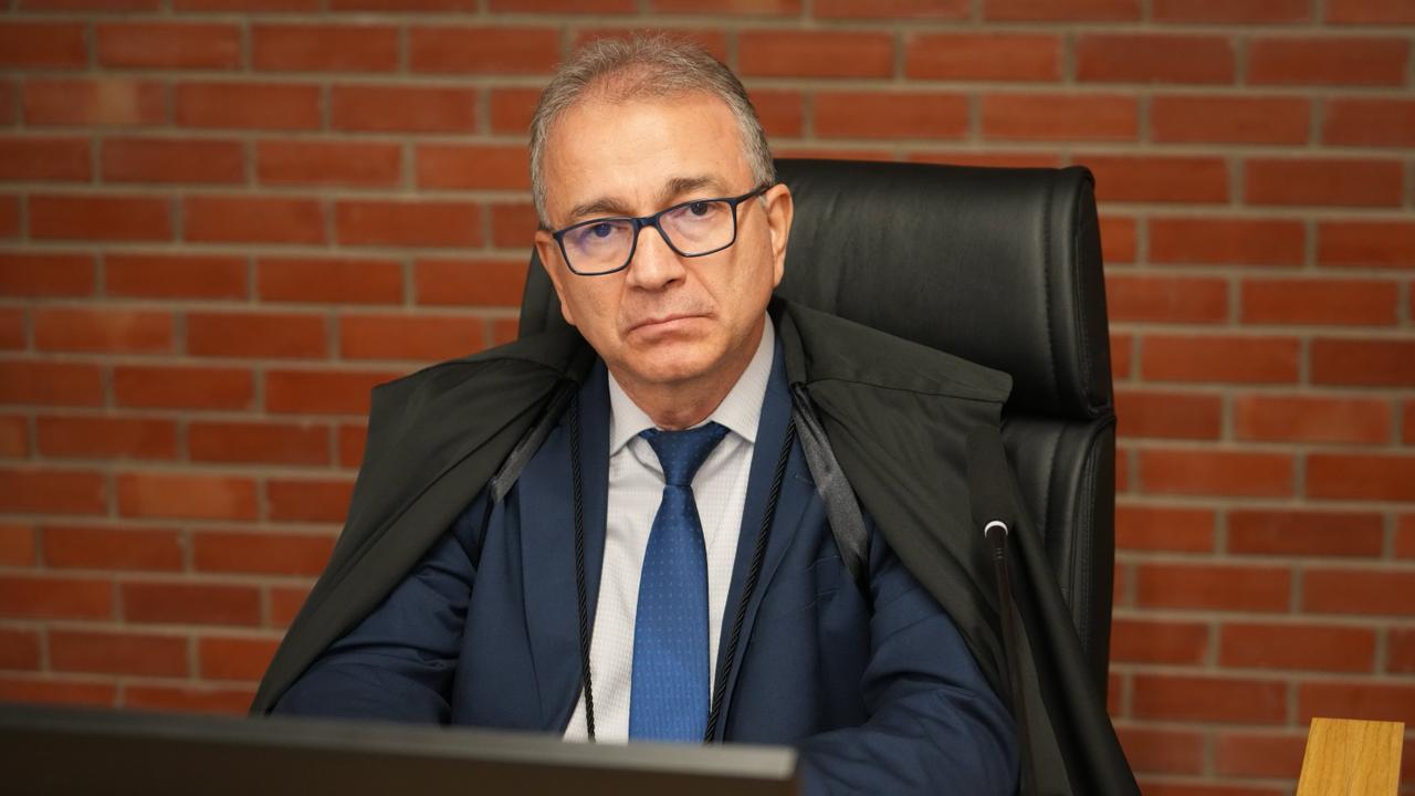 Desembargador Gilson Valadares, sentado em cadeira institucional, atrás de mesa de trabalho, em ambiente formal. Usa óculos, traje social escuro com gravata azul e toga sobre os ombros. Mantém postura ereta, com expressão séria, olhando à frente. À direita, há um microfone.