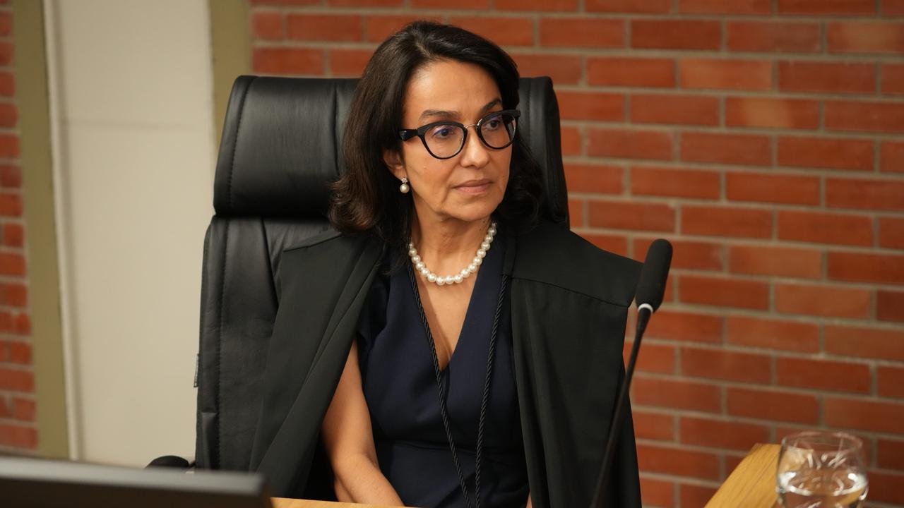 Desembargadora Hélvia Túlia, sentada em cadeira institucional, atrás de mesa de trabalho, em ambiente formal. Usa óculos, traje escuro e colar de pérolas, com toga sobre os ombros. Mantém postura ereta e olha para o lado. À frente, há um microfone e um copo com água. O fundo apresenta parede de tijolos aparentes.
