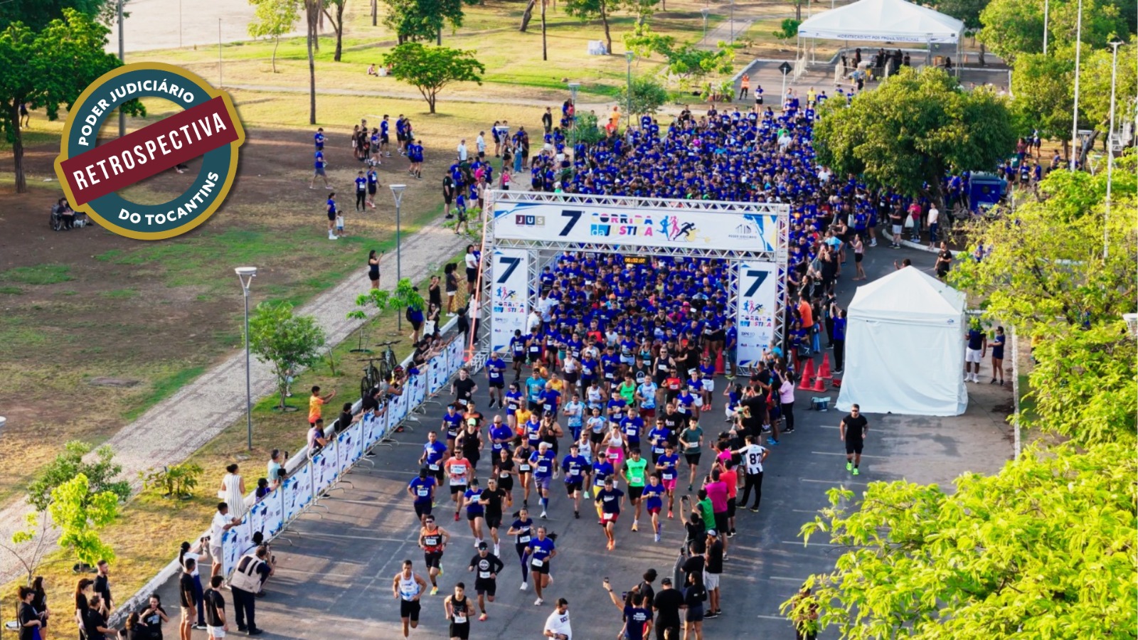A imagem mostra a 7ª Corrida da Justiça com centenas de corredores, muitos vestindo camisetas azuis, atravessam o pórtico de largada em uma via arborizada, enquanto espectadores acompanham o evento dos dois lados. No canto superior esquerdo, há um selo com a inscrição “Poder Judiciário do Tocantins – Retrospectiva”.