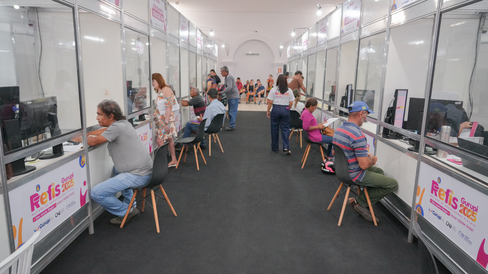 A foto mostra o mutirão do Refis Gurupi em um corredor com estandes de atendimento individualizados, equipados com computadores. Cidadãos estão sentados em frente aos balcões, realizando atendimentos, enquanto servidores circulam pelo espaço prestando orientações. Ao fundo, há pessoas aguardando em área de espera, evidenciando fluxo contínuo de atendimento em ambiente organizado e padronizado.