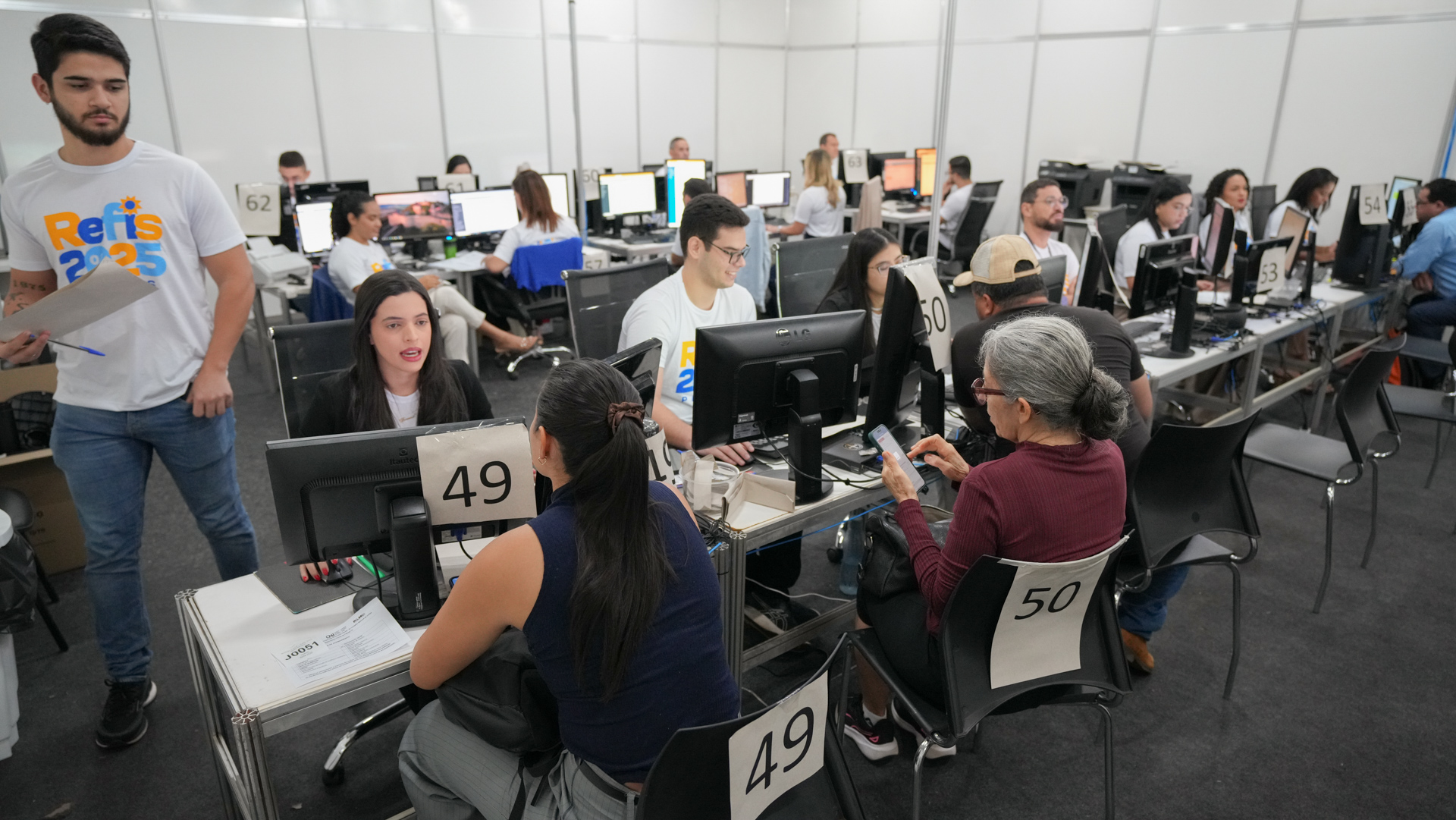 A foto mostra o mutirão do Refis em Palmas realizado em um ambiente amplo e organizado, com várias estações de atendimento numeradas, cada uma equipada com computador. Servidores, identificados com camisetas do Refis 2025, atendem cidadãos sentados à frente dos balcões, prestando orientações e realizando negociações. O espaço reúne diversas pessoas em atendimento simultâneo, evidenciando fluxo intenso, estrutura padronizada e atendimento individualizado ao público.
