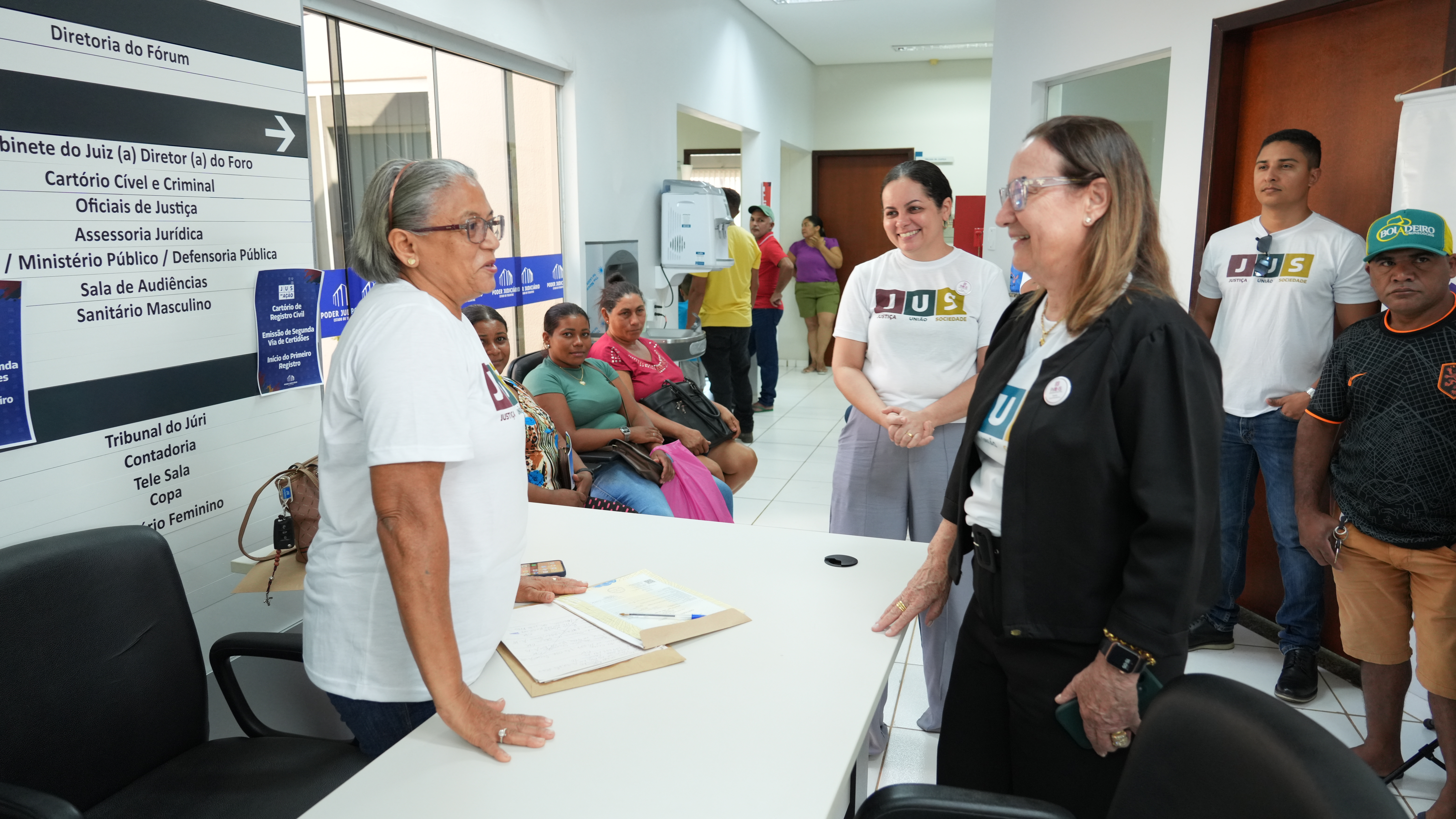 A foto mostra a presidente do TJTO cumprimentando servidores durante atendimento à população no âmbito do JUS, em espaço interno do fórum, com cidadãos aguardando e atividades em andamento.