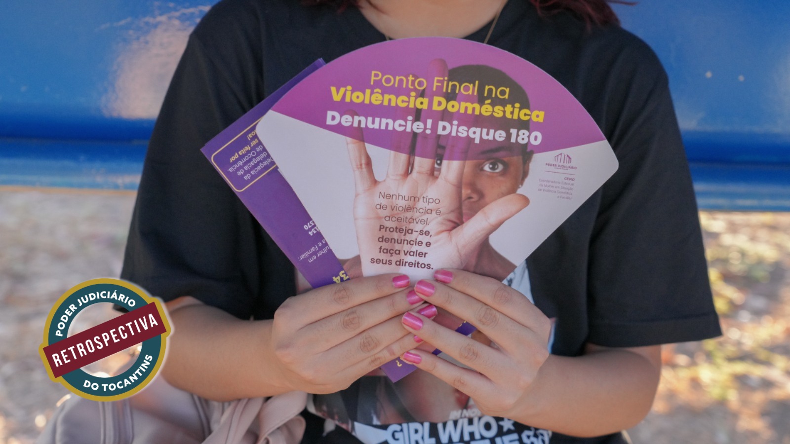 A imagem mostra uma pessoa segurando leques informativos de campanha contra a violência doméstica. O material traz a mensagem “Ponto final na violência doméstica. Denuncie! Disque 180” e destaca que nenhum tipo de violência é aceitável. O foco está na ação de conscientização e incentivo à denúncia, simbolizada pelo gesto de exibição do material educativo.