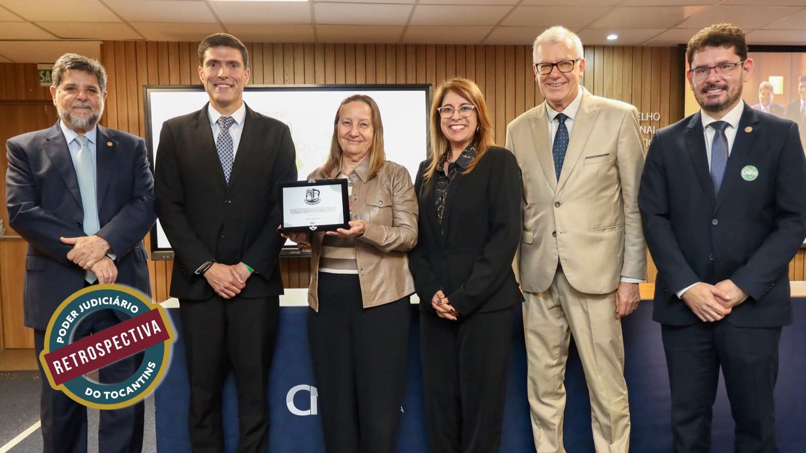 Registro de uma cerimônia institucional realizada em ambiente formal. Seis pessoas aparecem lado a lado, vestindo trajes sociais, em frente a uma mesa e a um painel com identidade institucional. No centro da imagem, uma mulher segura uma placa de homenagem, indicando o momento de reconhecimento ou premiação. Os participantes estão sorridentes, transmitindo um clima de solenidade e celebração, típico de eventos oficiais de entrega de honrarias.