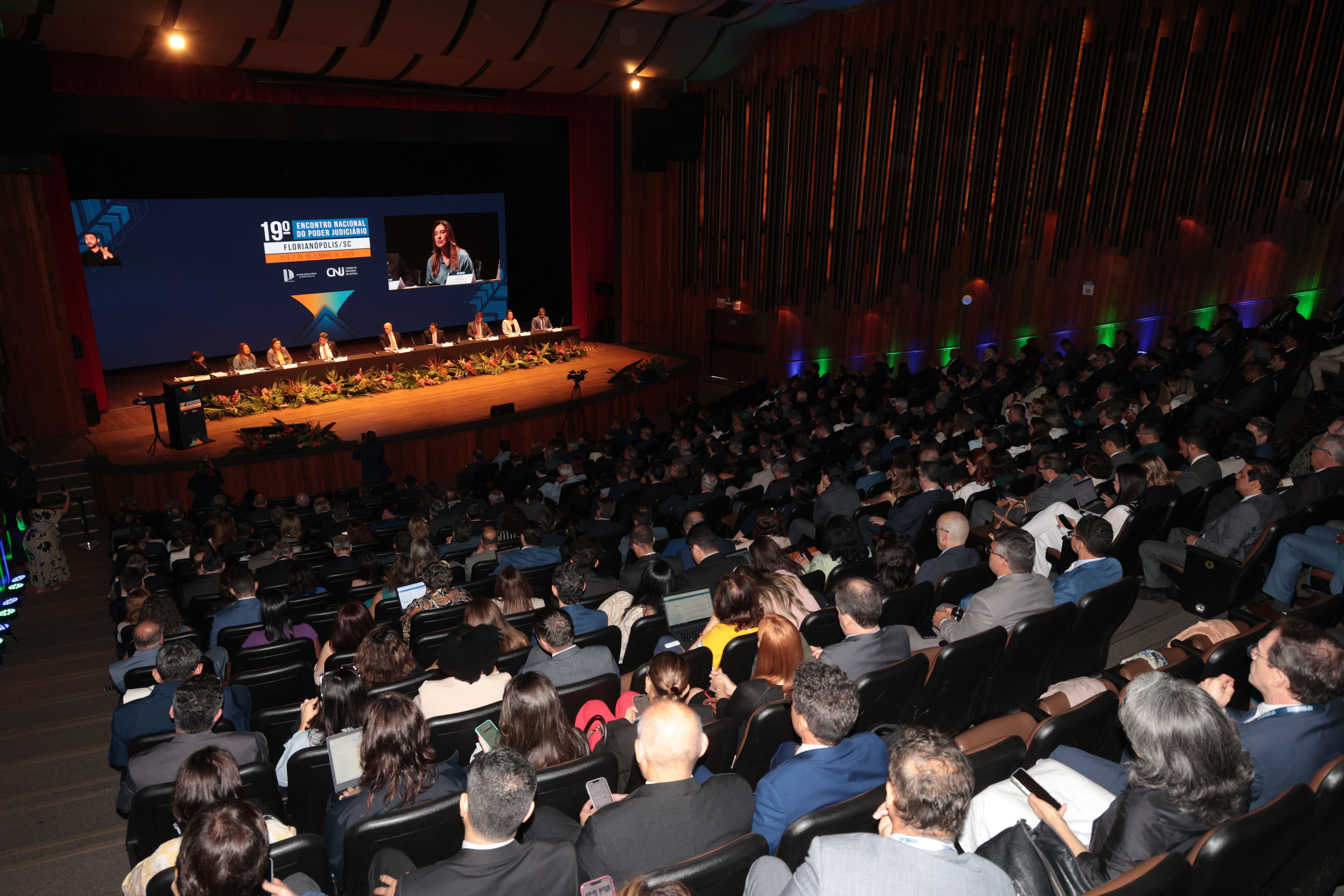 Auditório lotado com plateia assistindo a uma mesa de autoridades no palco durante o 19º Encontro Nacional do Poder Judiciário, em Florianópolis (SC). Um grande telão ao fundo exibe a imagem de uma palestrante