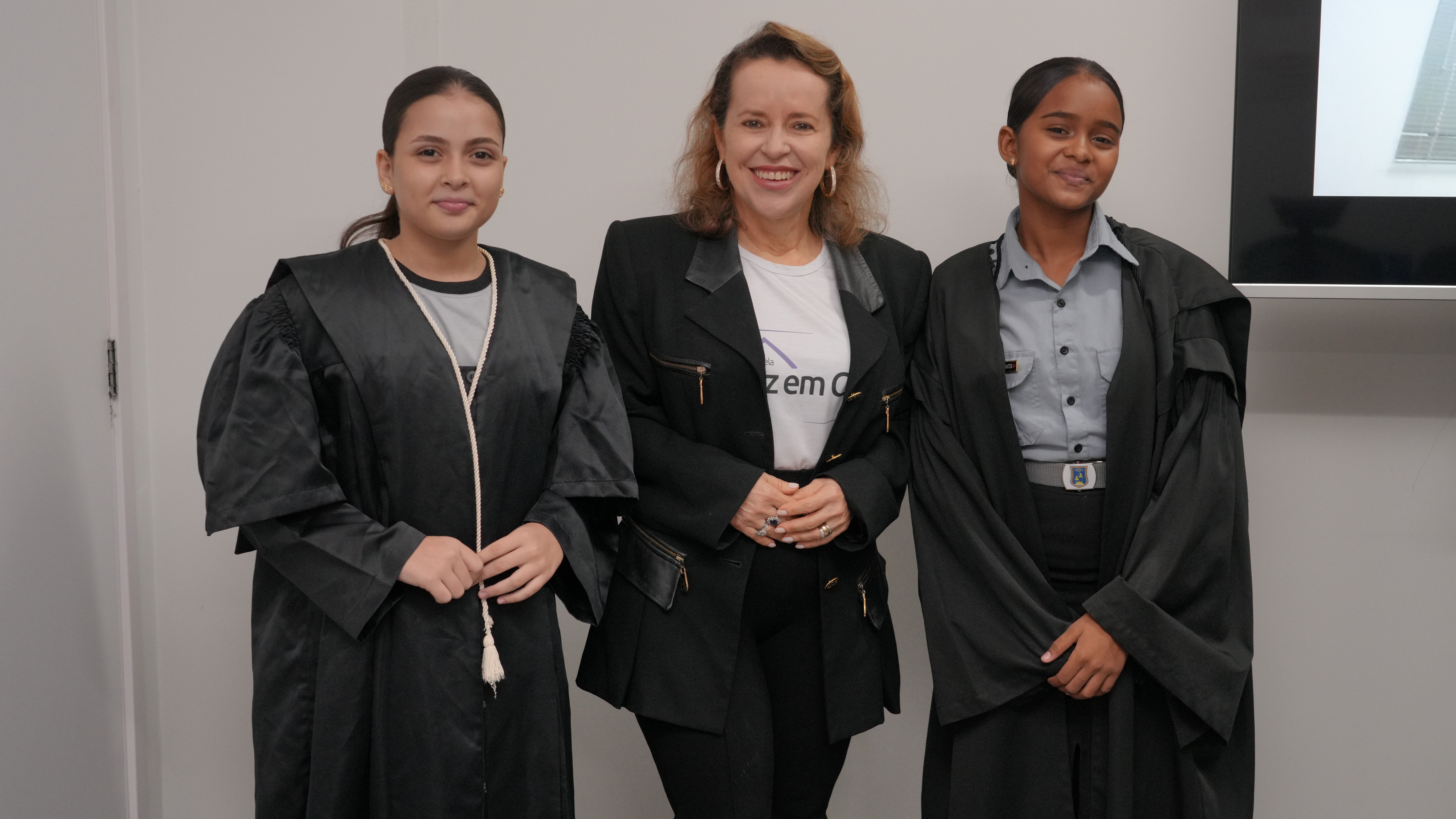 Três mulheres posam sorrindo para a foto. As duas jovens das laterais vestem togas e a mulher ao centro usa blazer preto e camiseta branca. Elas estão em um ambiente interno, com parede clara ao fundo