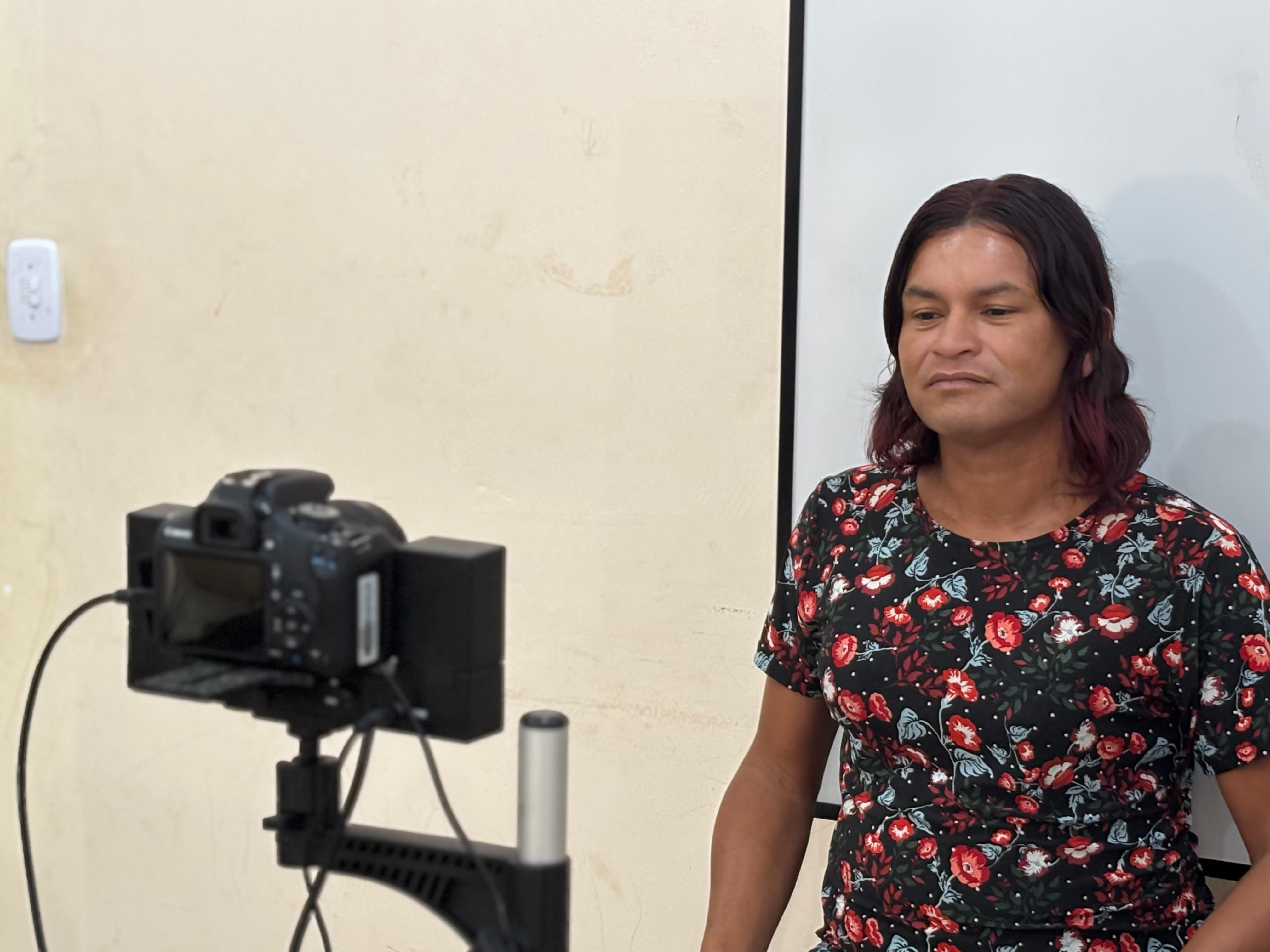 A foto mostra uma pessoa sentada em frente a uma câmera fotográfica montada em suporte, como parte de um procedimento de registro. Ela veste uma blusa floral escura e está posicionada diante de uma parede clara com um quadro branco ao lado. A câmera está voltada diretamente para ela, capturando sua imagem.