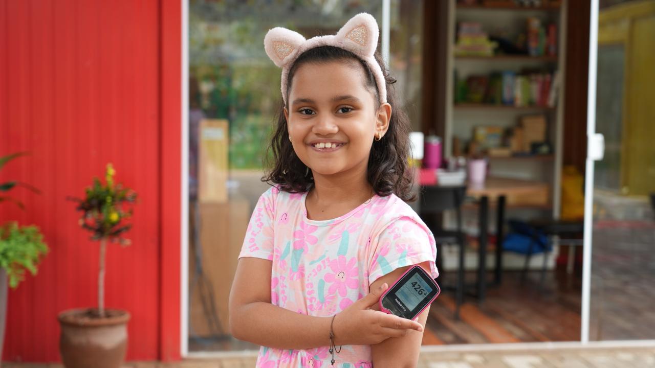 A foto mostra a menina Maria Tereza sorrindo, usando tiara de orelhas de gato e vestido rosa estampado. Ela exibe um monitor de glicemia apoiado no braço, onde também há um sensor aplicado. Ao fundo, aparecem uma parede vermelha, plantas em vasos e um ambiente interno com prateleiras e mesas.