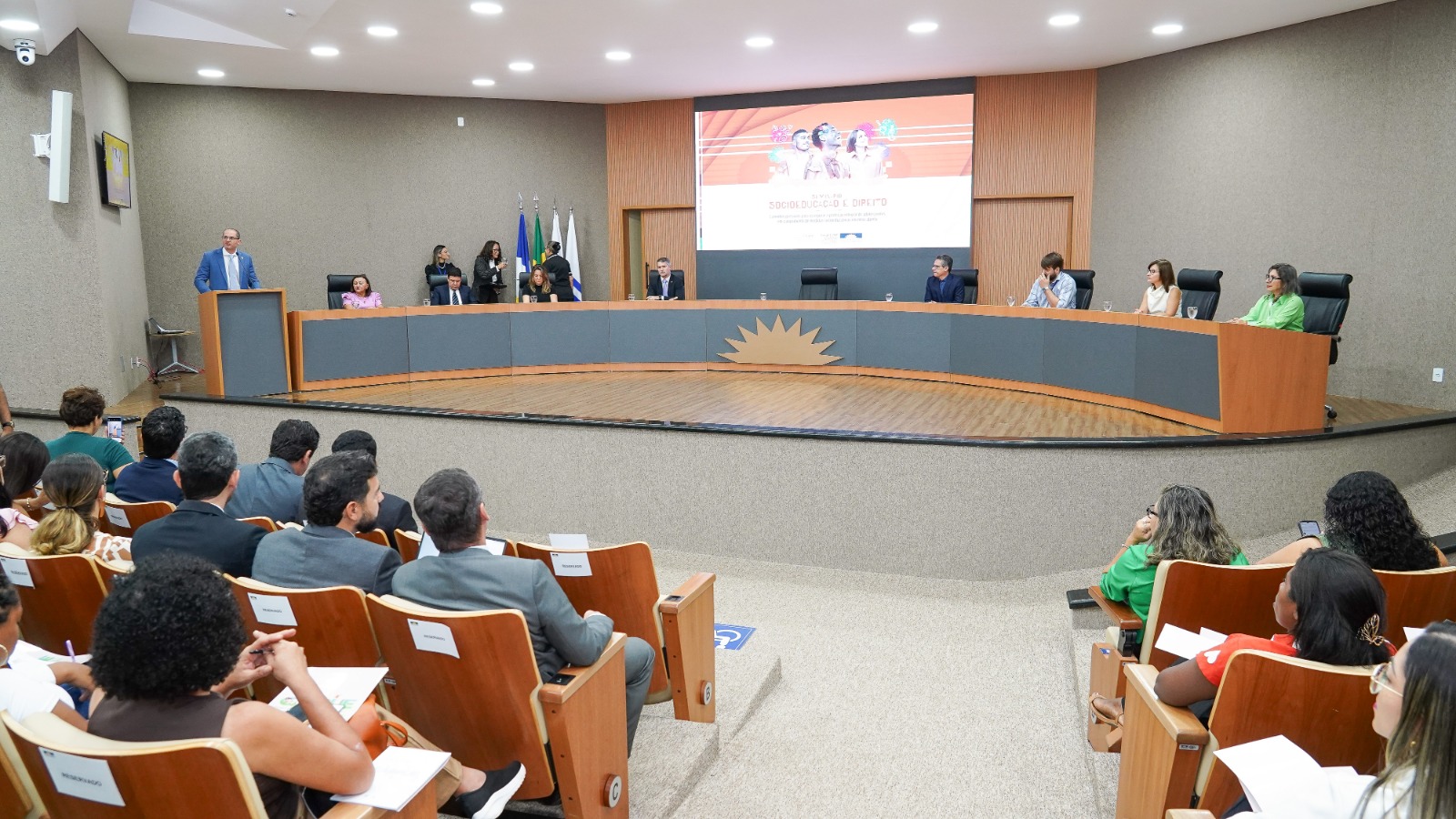 A imagem mostra um auditório com várias pessoas sentadas em cadeiras em fileiras e um grupo de autoridades sentadas atrás de uma mesa no palco. Na cena aparece também um homem em pé, posicionado atrás de um púlpito discursando.