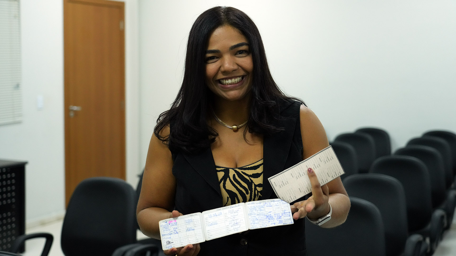 A imagem mostra uma mulher sorridente de cabelos escuros e lisos, vestindo colete preto e blusa com estampa animal print. Ela está em uma sala com cadeiras de auditório ao fundo, segurando uma caderneta de vacinação aberta em uma mão e um cartão de vacinação na outra. Ao fundo, é possível ver uma porta de madeira fechada e parte de uma persiana branca. A expressão da mulher transmite simpatia e satisfação.
