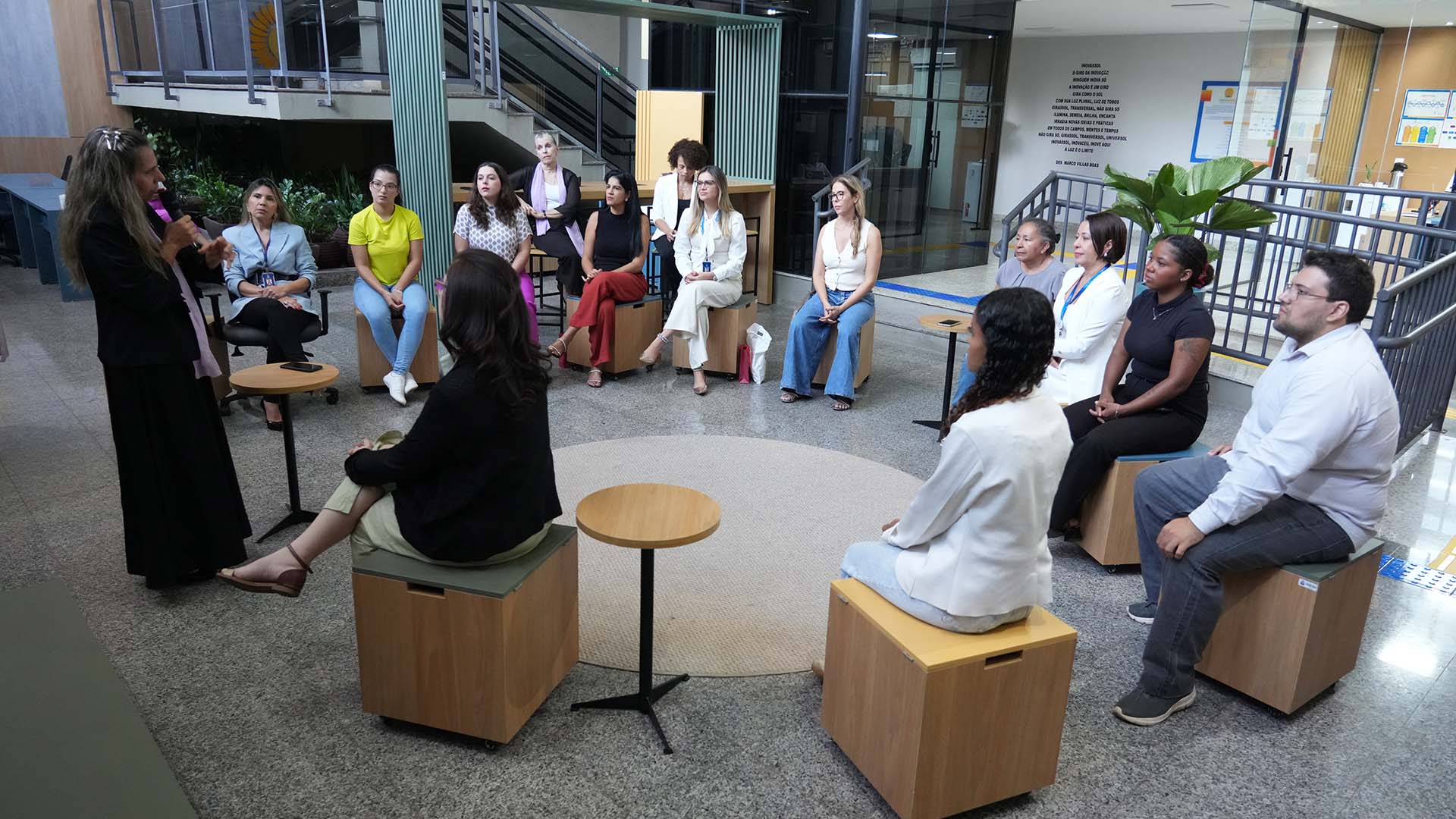 A foto mostra um grupo de mulheres reunidas em um espaço interno, organizado em forma de roda. Todas estão sentadas em bancos individuais de madeira, escutando atentamente uma mulher que fala em pé, segurando um microfone.