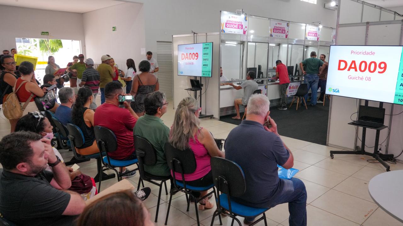 Fotografia colorida onde várias pessoas estão sentadas em cadeiras azuis em uma ampla sala fechada enquanto esperam atendimento. Nos cantos esquerdo e direito duas televisões mostram as senhas que foram distribuídas para as pessoas sentadas nas cadeiras. Em meio as televisões, um corredor se abre onde as pessoas estão sendo direcionadas para o atendimento. Algumas pessoa inclusive estão sendo atendidas. 