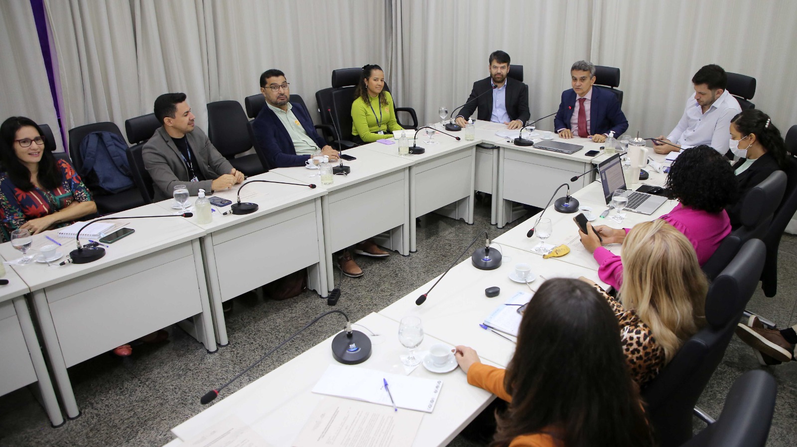Fotografia colorida onde um grupo de homens e mulheres estão sentados em volta de uma mesa retangular que possui uma grande abertura no meio. Eles estão em uma sala branca com uma cortina também branca de fundo