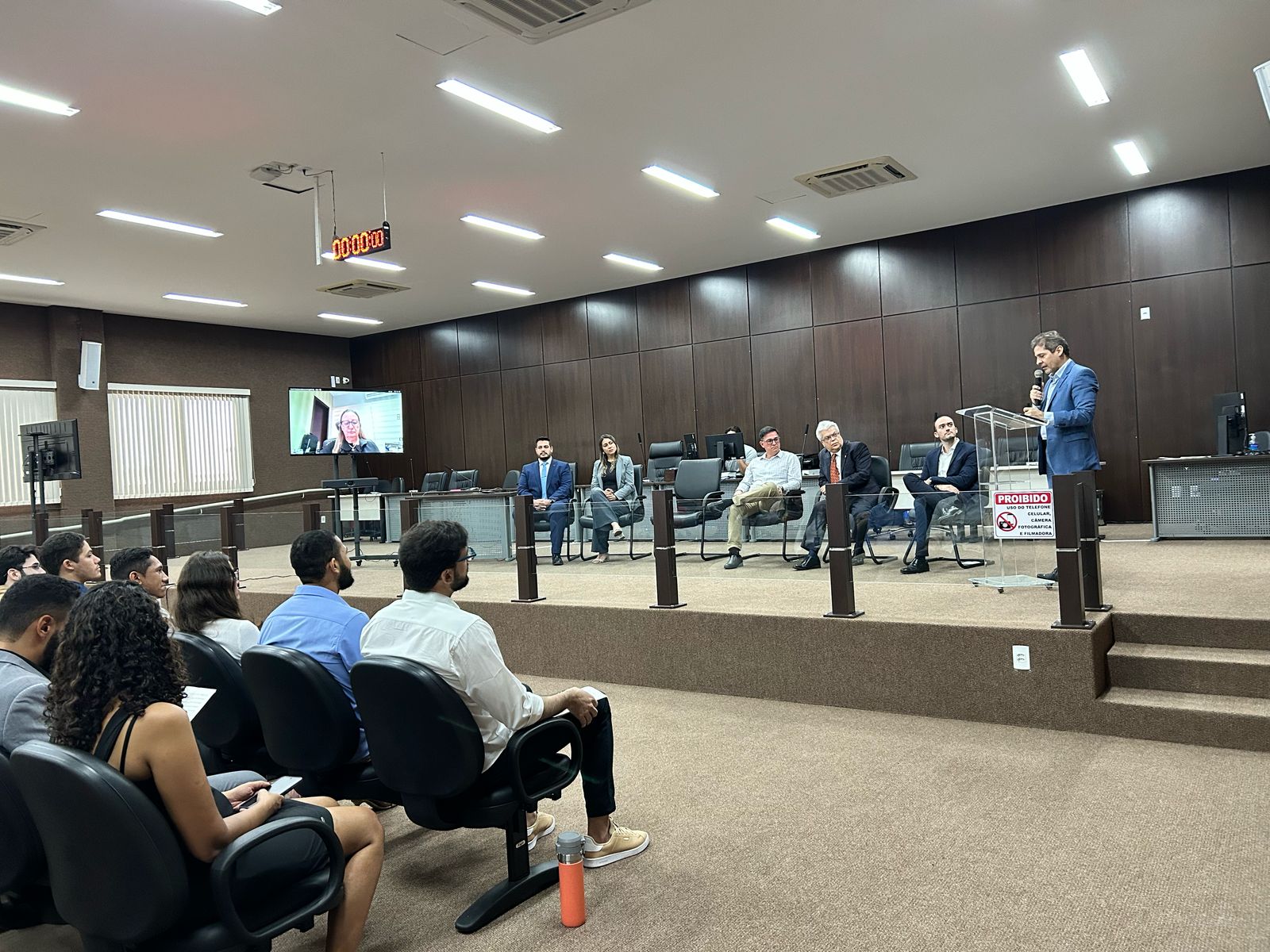 Fotografia colorida onde várias pessoas estão sentadas em cadeiras pretas em um auditório fechado. Em frente ao grupo, sete pessoas estão sentadas em uma área mais alta, também sentadas em cadeiras. Do lado esquerdo, uma televisão com a imagem de uma mulher vestida de preto com óculos e do lado direito, um homem de roupa social atrás de um púlpito. Atrás deles uma parede marrom e um telhado branco acima.  