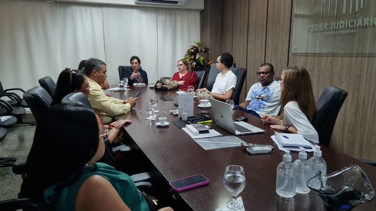 Fotografia colorida onde um grupo de nove pessoas estão sentadas em volta de uma longa mesa de madeira escura retangular. Elas estão em reunião, e em cima da mesa estão dispostos diversos objetos. 