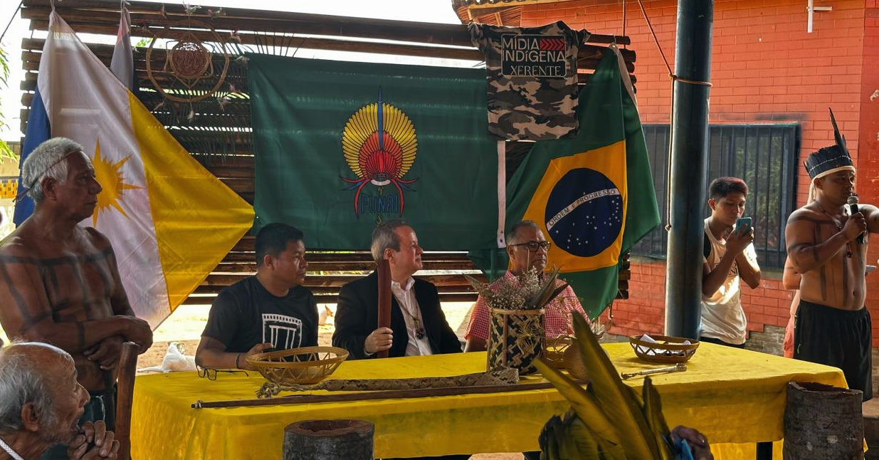 Imagem mostra mesa com indígenas e não indígenas durante reunião na aldeia xerente em Tocantinia e ao fundo um painel de madeira com bandeira do Brasil, da Funai e artesanato indígena