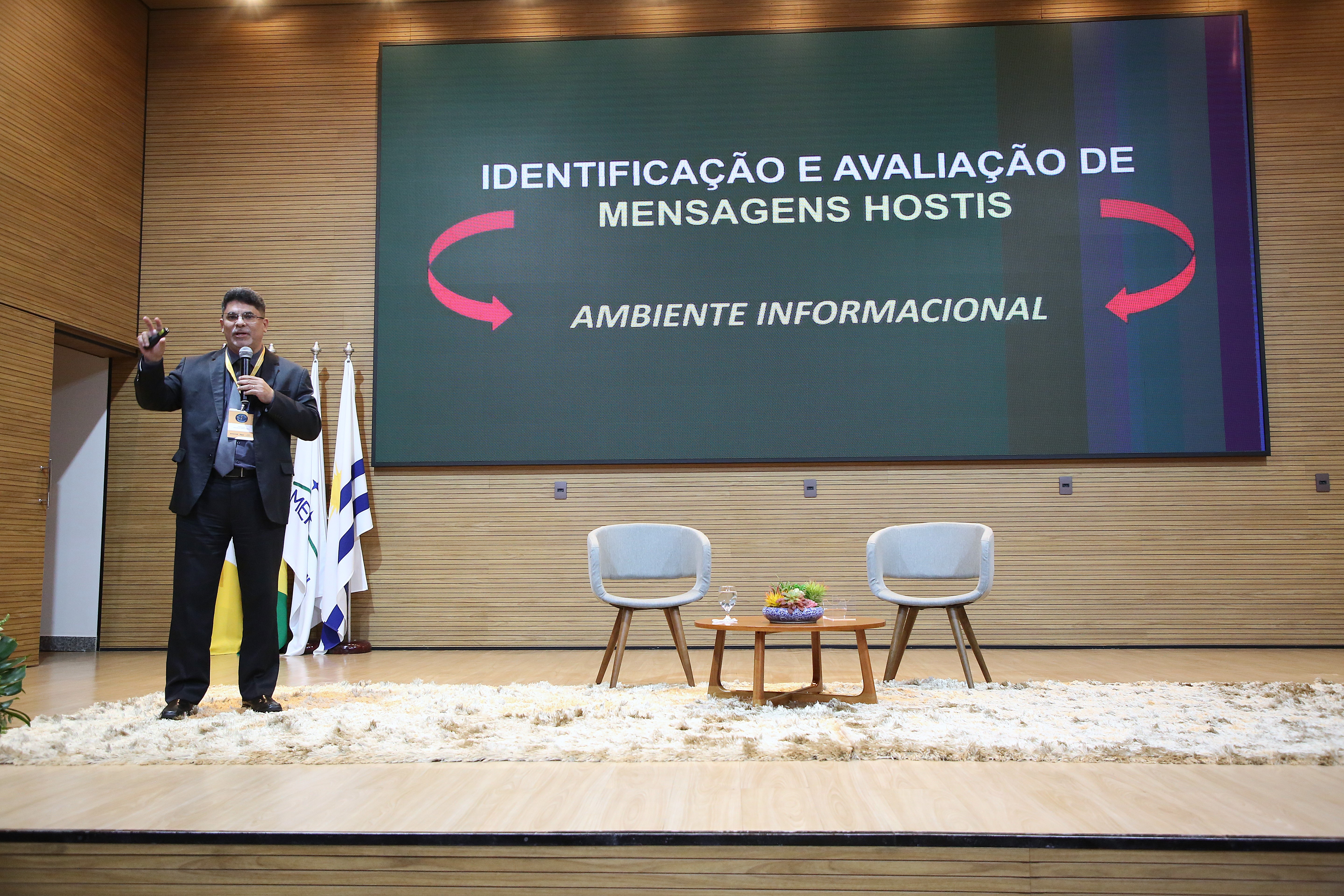  Imagem mostra o palestrante,  de terno escuro, no palco do auditório, ministrando a palestra. Em frente a uma tela grande que exibe informações sobre o evento. Os elementos estão dispostos de forma a direcionar o foco para o palestrante. O palestrante está de pé no palco, em frente à tela. Há duas cadeiras e uma mesinha, vazias, à frente da tela, em ambos os lados. 