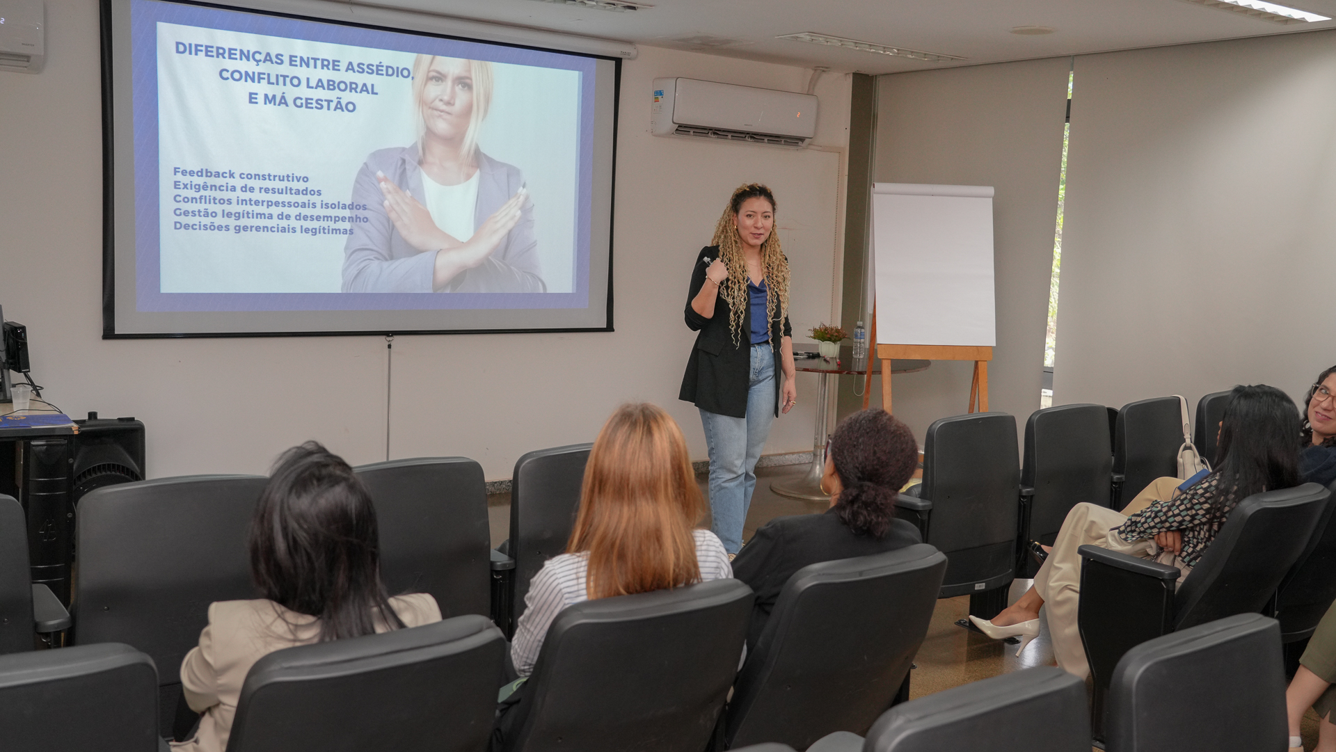 Imagem mostra pessoas sentadas no auditório da Controladoria Geral do Estado diante de uma mulher falando, Tatiane Dias, durante whorkshop sobre assedio 