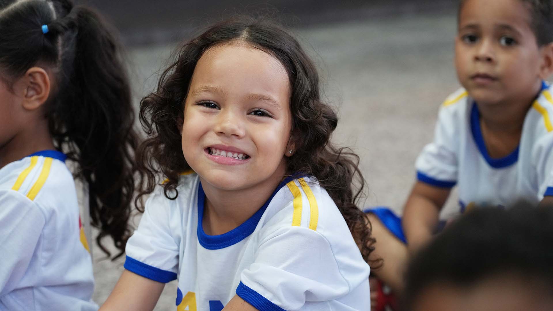 Foto de uma criança sorrindo; ela aparece vestida com o uniforma da escola nas cores branca e azul