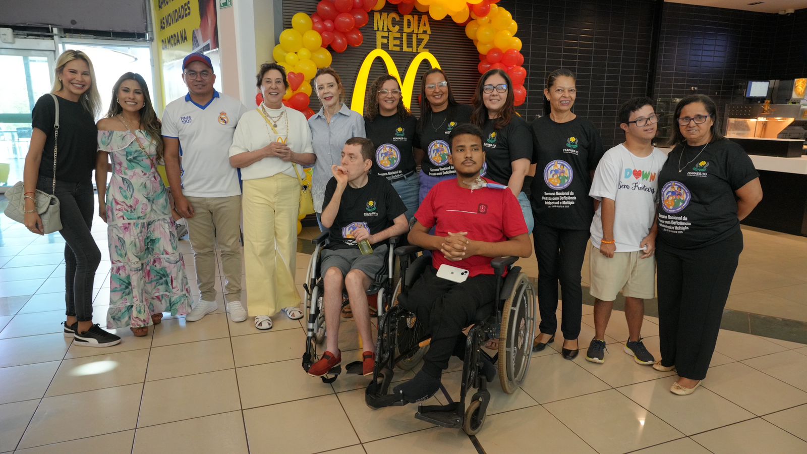 Grupo de treze pessoas posando em frente a uma decoração de balões vermelhos e amarelos em formato de arco, com a inscrição 'McDia Feliz' no centro. Todos sorriem, segurando sacolas e caixas de lanche, em clima de confraternização e apoio à causa social do evento