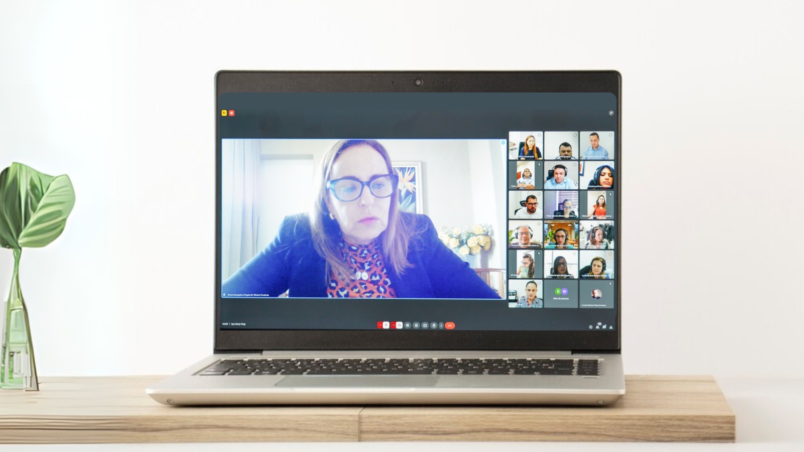 A imagem mostra uma tela de computador com uma videoconferência em andamento. A mulher no centro da tela do laptop está em uma reunião on-line com vários outros participantes. Há uma planta pequena ao lado do laptop.