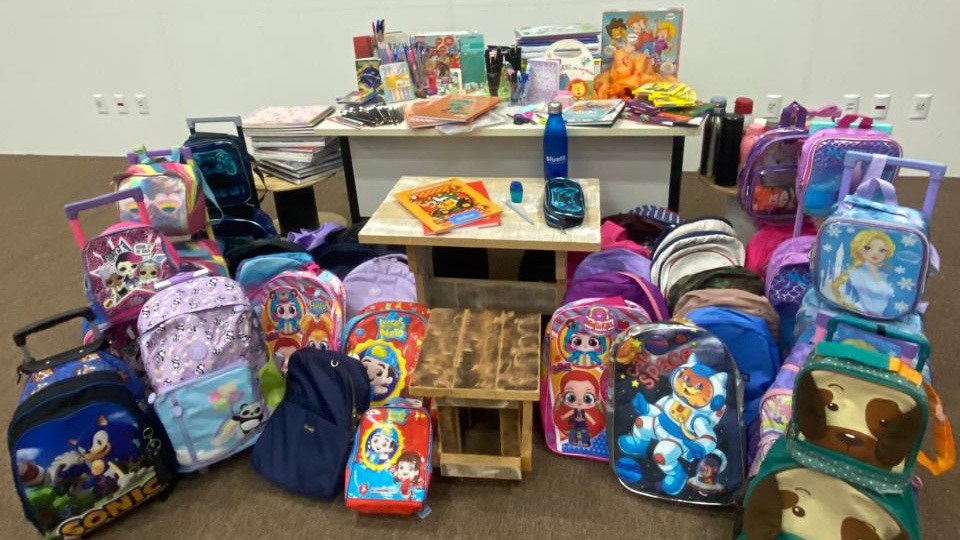 Mochilas, estojos, livros e materiais escolares novos e usados organizados em uma sala, dispostos sobre mesas e no chão, como parte de uma campanha de arrecadação para estudantes