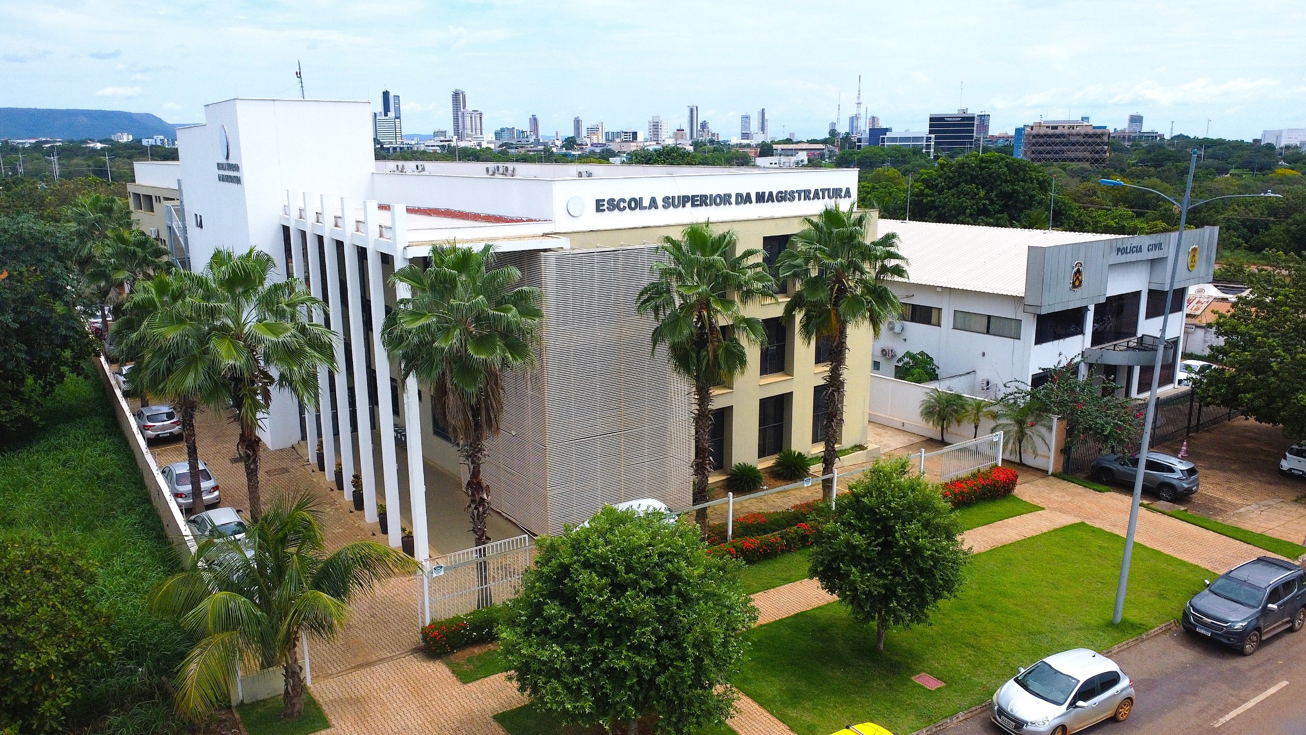 Vista aérea da fachada da Escola Superior da Magistratura, com prédio moderno em tons claros, colunas verticais, palmeiras no entorno, área verde bem cuidada e edificações institucionais ao lado, em uma região urbana arborizada
