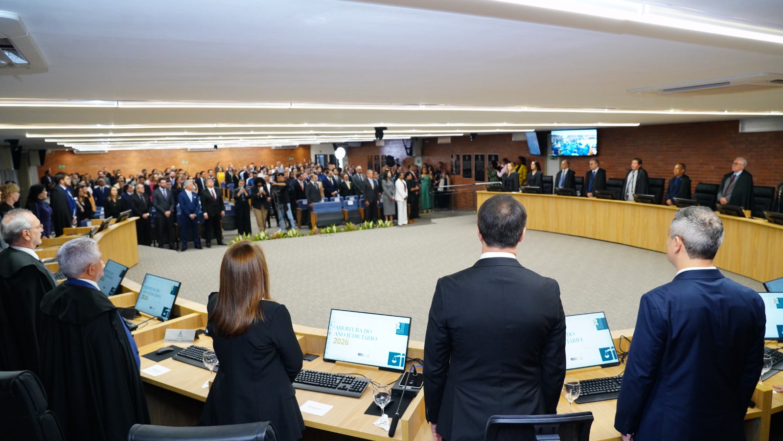Vista ampla de sessão solene em plenário judicial, com magistrados e autoridades de pé à mesa diretiva, público em pé no auditório, computadores sobre a bancada e arranjos florais ao centro do espaço. Ambiente interno institucional.