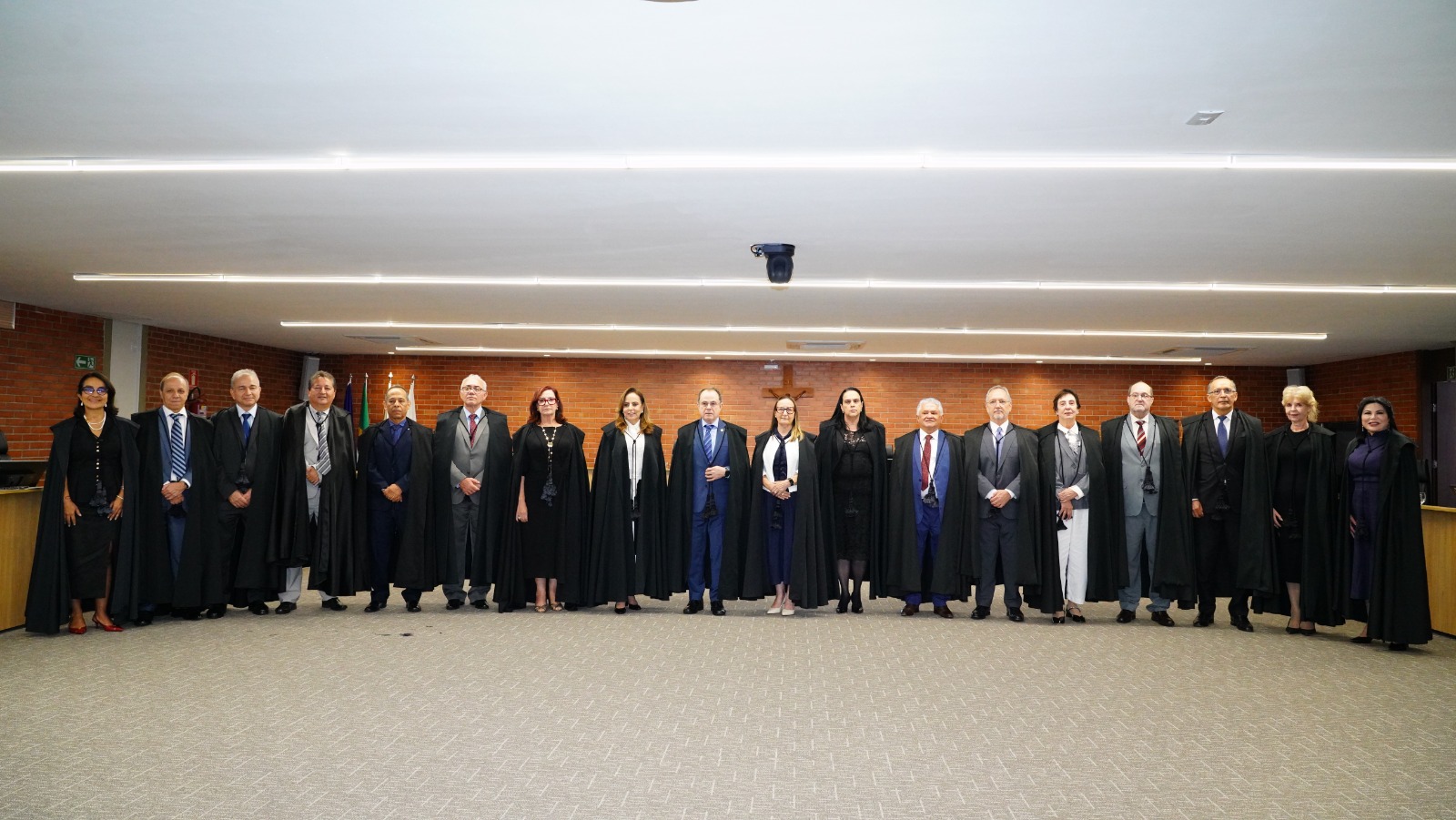 Foto oficial com magistrados e magistradas em trajes de toga, alinhados lado a lado no plenário judicial, em ambiente interno institucional, com parede de tijolos ao fundo e iluminação no teto.