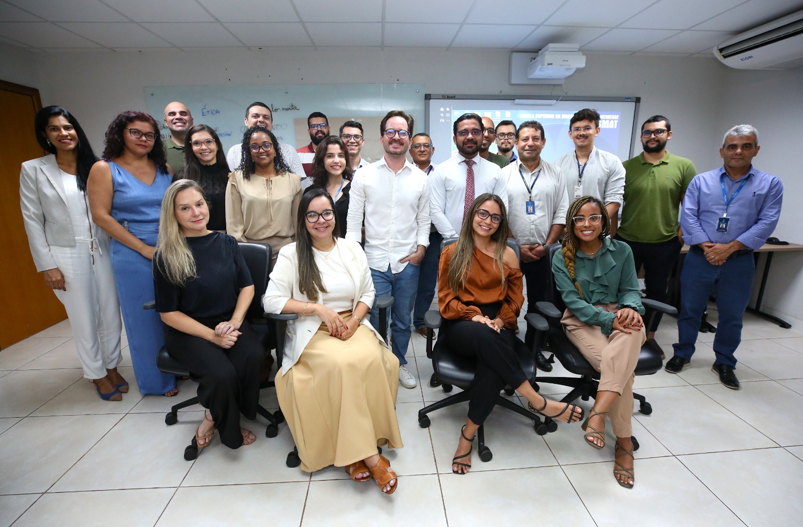 A imagem mostra um grupo de profissionais reunidos em um ambiente corporativo, aparentemente uma sala de reuniões ou treinamento. Ao todo, cerca de 18 pessoas posam para a foto, distribuídas entre a primeira fila sentada e os demais em pé, ao fundo.  Na primeira fila, quatro mulheres estão sentadas em cadeiras giratórias pretas. Elas vestem trajes sociais em tons neutros e terrosos, como preto, bege, marrom e verde, transmitindo elegância e formalidade. Todas sorriem para a câmera.  Atrás delas, homens e mulheres estão posicionados lado a lado, também trajando roupas sociais ou esporte fino — camisas sociais, blazers, vestidos e calças sociais. Alguns utilizam crachás de identificação, reforçando o caráter institucional do encontro. O grupo demonstra um clima de integração e cordialidade.