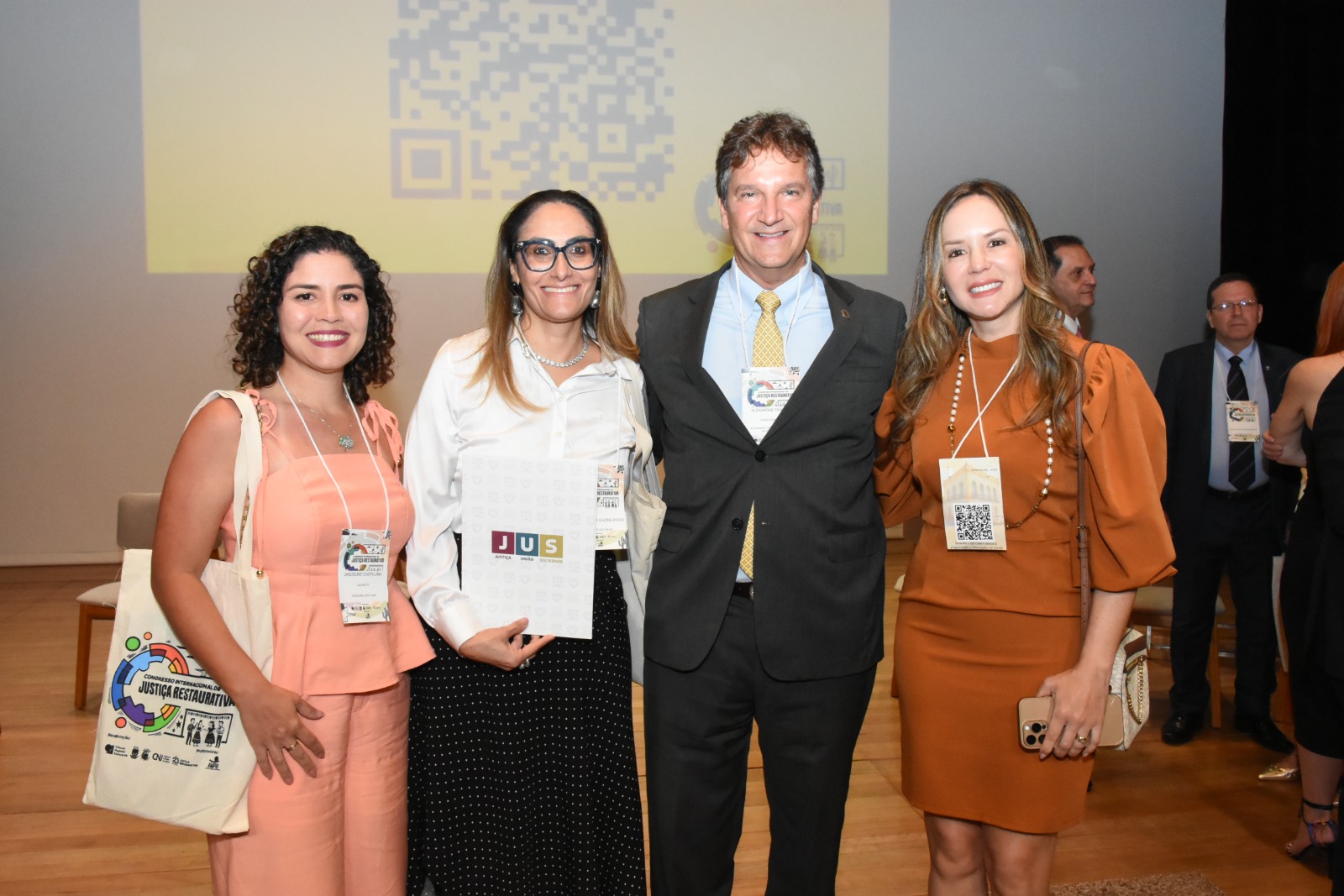 Quatro participantes posam para foto durante o congresso de Justiça Restaurativa. Duas mulheres e um homem vestem trajes formais e utilizam credenciais do evento; ao centro, o homem está de terno escuro e gravata amarela. Uma das participantes segura uma pasta com a identidade visual do congresso, enquanto outra traz uma bolsa temática do evento.  Ao fundo, há a projeção de um QR code na tela do auditório, reforçando o ambiente institucional e acadêmico da ocasião.