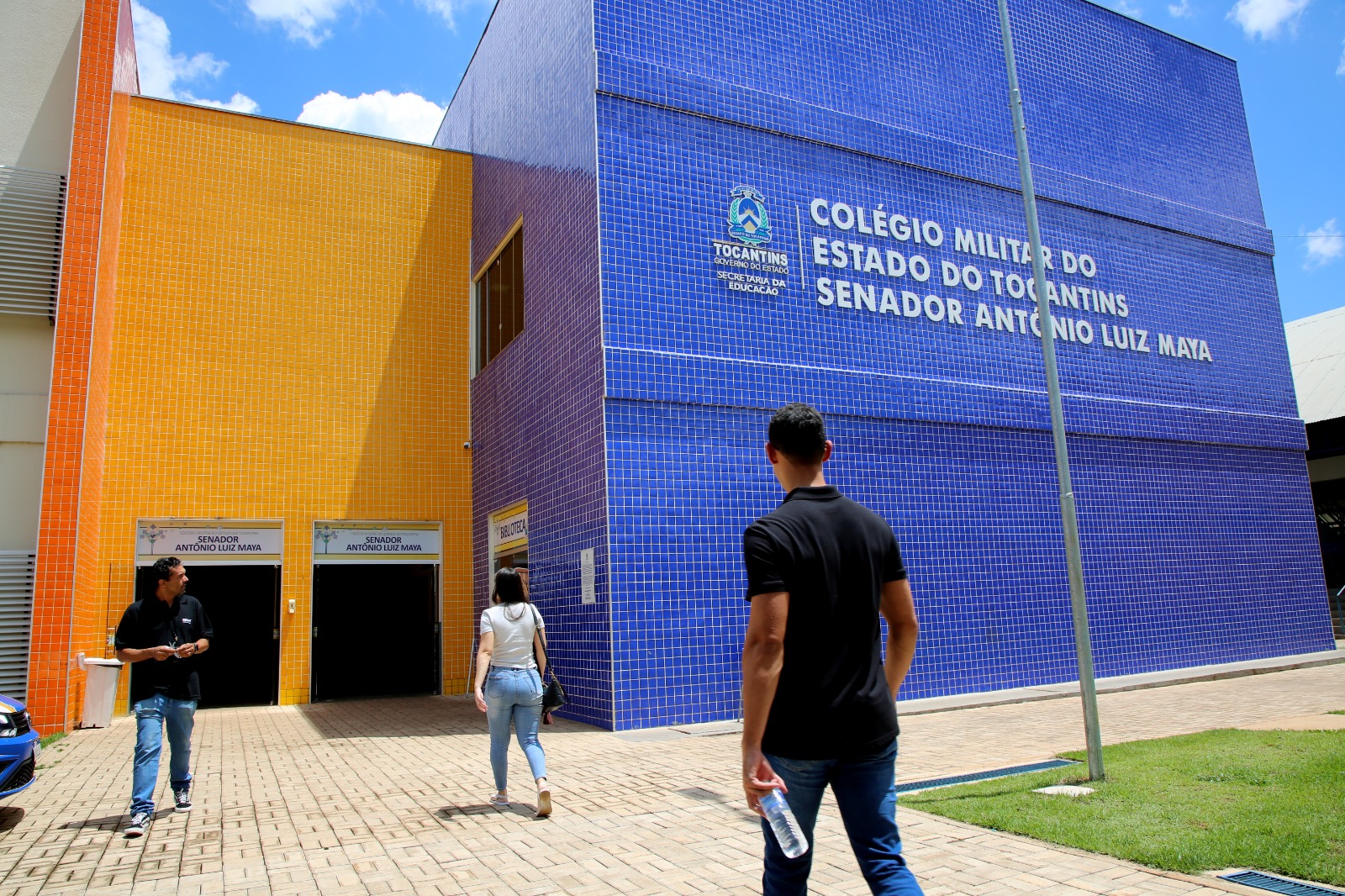 A imagem mostra a fachada do Colégio Militar do Estado do Tocantins – Senador Antônio Luiz Maya, com paredes em azulejos azuis e laranja e o nome da instituição em destaque. À frente do prédio, três pessoas caminham em direção à entrada, em uma área externa com piso de blocos e gramado sob céu parcialmente nublado.