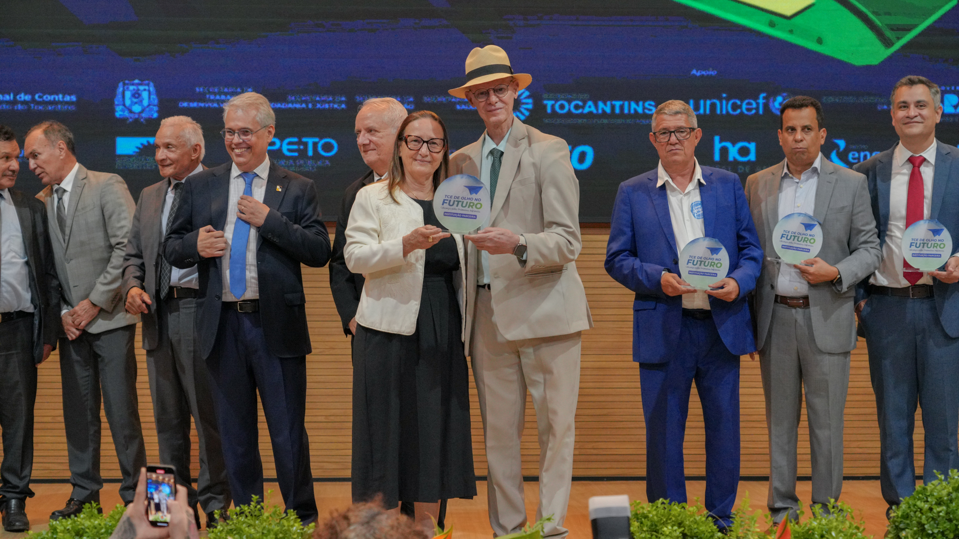 No palco do auditório do TJTO, autoridades e representantes de instituições públicas posam lado a lado durante uma cerimônia de reconhecimento. Ao centro, presidente do TJTO e presidente do TCETO seguram juntos uma placa circular referente ao Selo de Menção Honrosa do projeto “TCE de Olho no Futuro”, enquanto outros participantes também exibem placas semelhantes
