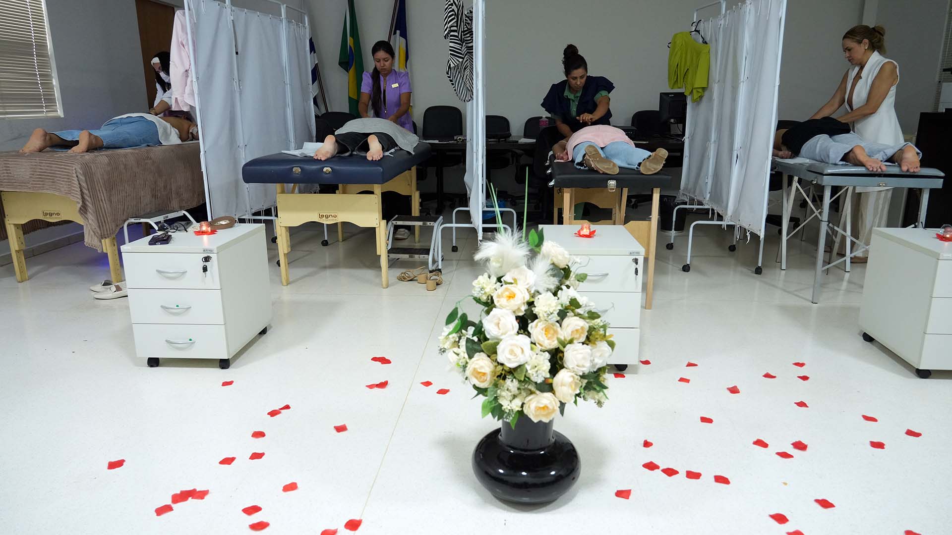 Quatro pessoas estão deitadas em macas enquanto profissionais realizam massagens terapêuticas. As macas são separadas por cortinas brancas, garantindo privacidade. Ao centro do espaço há um arranjo de flores brancas em vaso preto e pétalas vermelhas espalhadas pelo chão