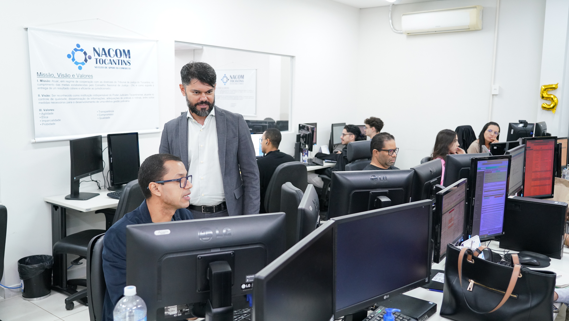Ambiente de trabalho do NACOM Tocantins, com servidores atuando em computadores. Em destaque, o juiz Wellington Magalhães, coordenador do NACOM, acompanha e orienta as atividades da equipe no setor. Ao fundo, banner institucional do núcleo na parede.