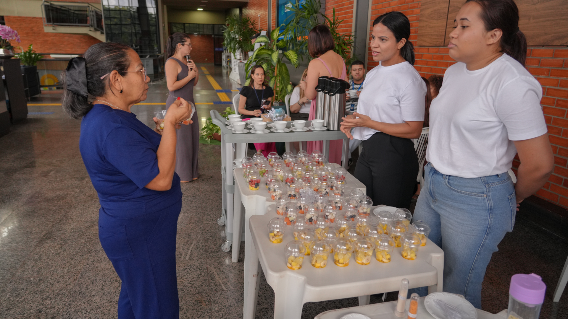 A imagem mostra servidoras em um espaço interno do prédio do Tribunal de Justiça do Estado do Tocantins participando de uma atividade de degustação de frutas durante ação de saúde e nutrição. Sobre uma mesa estão diversos copos com porções de frutas, enquanto duas organizadoras conversam com uma participante sobre a experiência alimentar proposta.