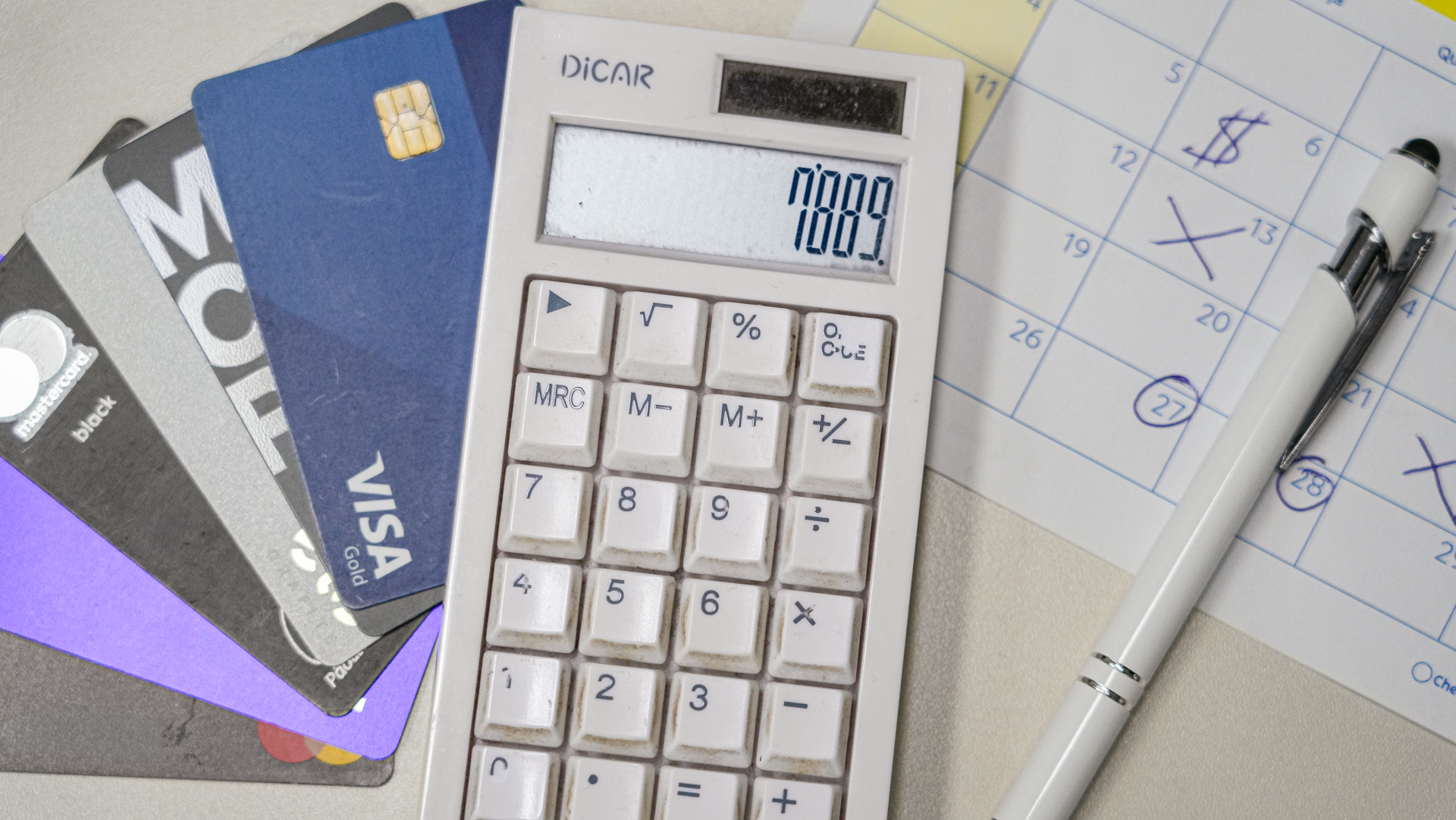 Imagem de uma calculadora sobre uma mesa, exibindo números no visor, ao lado de vários cartões de crédito parcialmente sobrepostos. À direita, há uma agenda com datas marcadas e anotações, acompanhada de uma caneta.