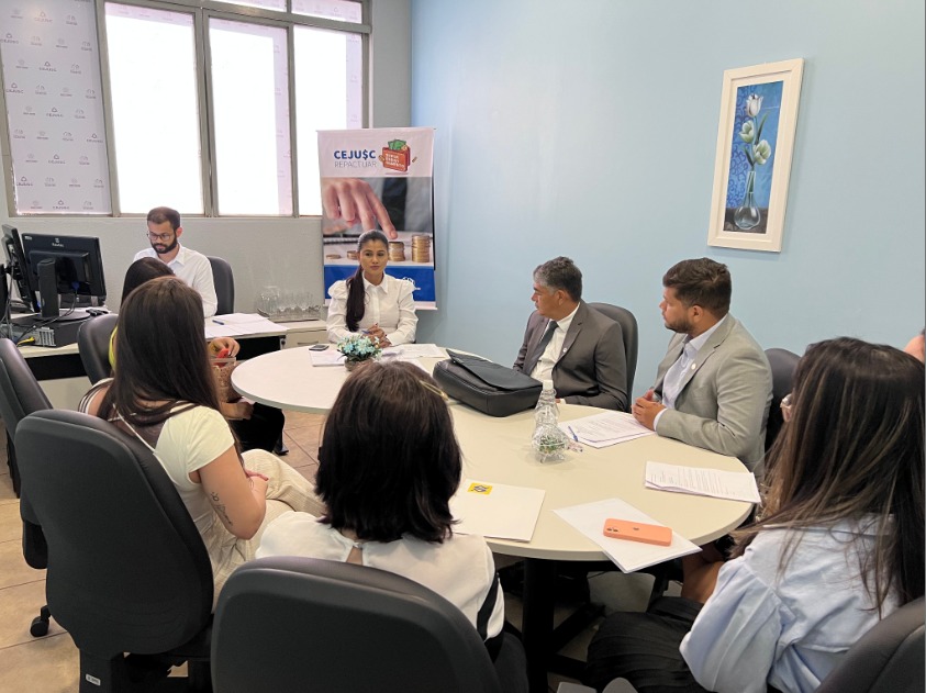 Reunião de atendimento no CEJUSC, com equipe e participantes reunidos em torno de uma mesa redonda durante uma audiência de conciliação. Ao centro, uma mediadora conduz o diálogo, enquanto os demais acompanham com documentos em mãos. Ao fundo, banner do programa CEJUSC Repactuar reforça o foco na reorganização financeira.