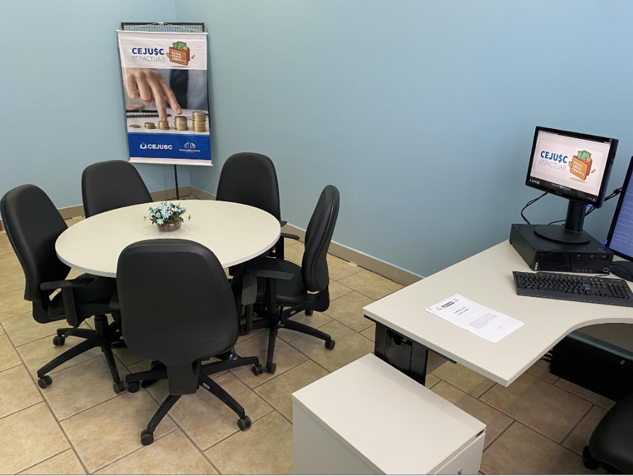 Ambiente interno de atendimento do CEJUSC, com mesa redonda ao centro cercada por cadeiras, preparada para reuniões ou audiências de conciliação. Ao fundo, um banner do programa CEJUSC Repactuar com imagem de moedas empilhadas, remetendo à organização financeira. À direita, há uma mesa de trabalho com computador, teclado e documentos, compondo um espaço funcional e acolhedor voltado ao atendimento do público.