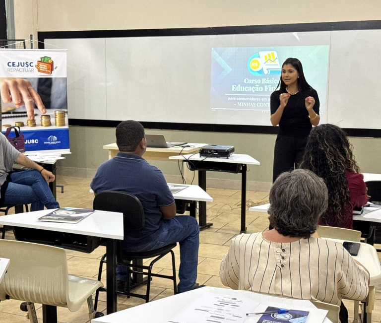 Sala de aula com atividade do programa CEJUSC Repactuar, onde uma instrutora conduz o curso de educação financeira “Minhas Contas em Dia”. Participantes estão sentados, atentos à apresentação exibida em projetor, que aborda organização das finanças pessoais. Ao lado, um banner do programa reforça a proposta de orientação e renegociação de dívidas.