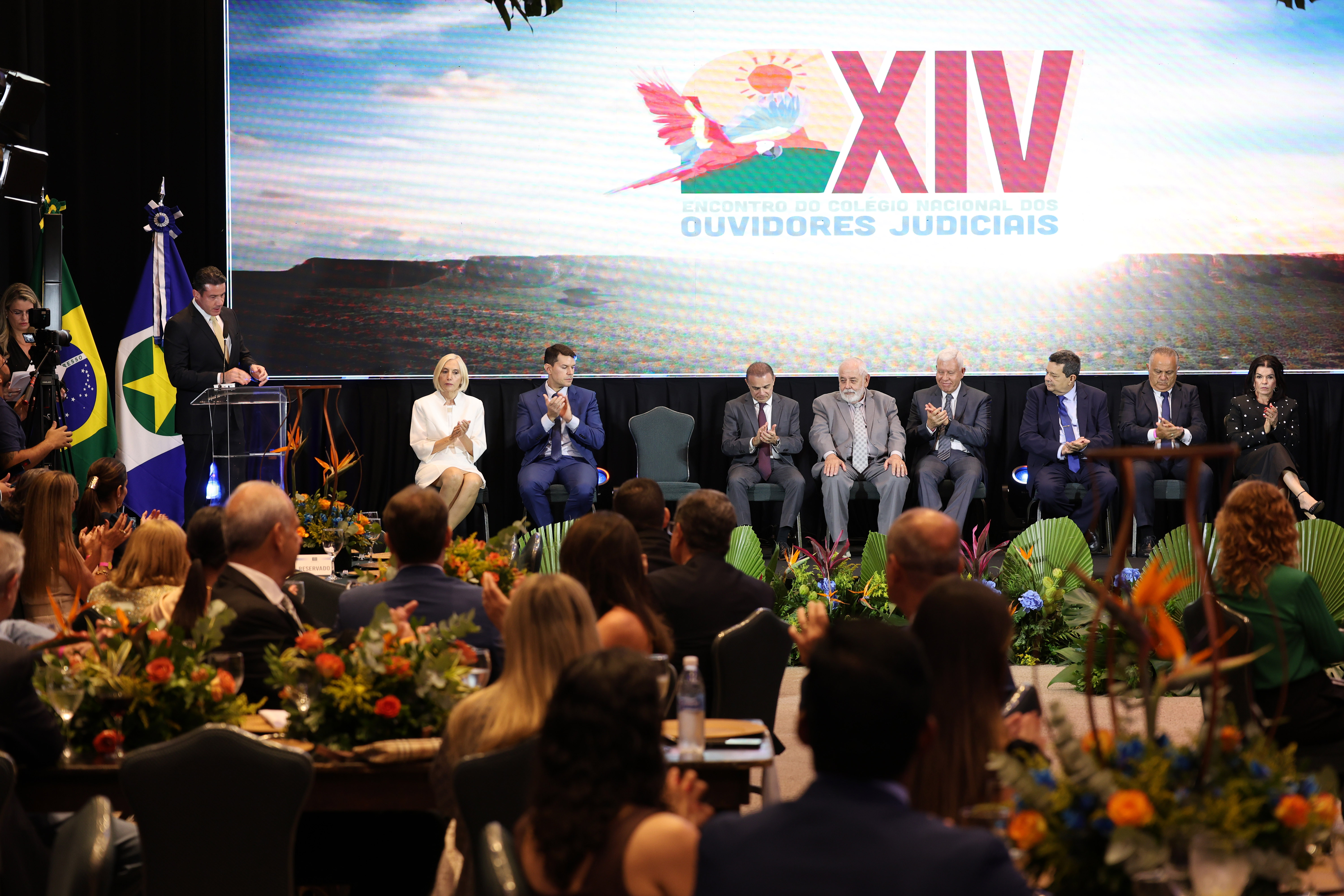 Solenidade de abertura de evento institucional com autoridades sentadas em palco. À esquerda, um homem discursa em púlpito ao lado da bandeira. Ao fundo, telão exibe a identidade visual do XIV Encontro do Colégio Nacional dos Ouvidores Judiciais. Em frente ao palco, há decoração com plantas ornamentais e participantes ao público.