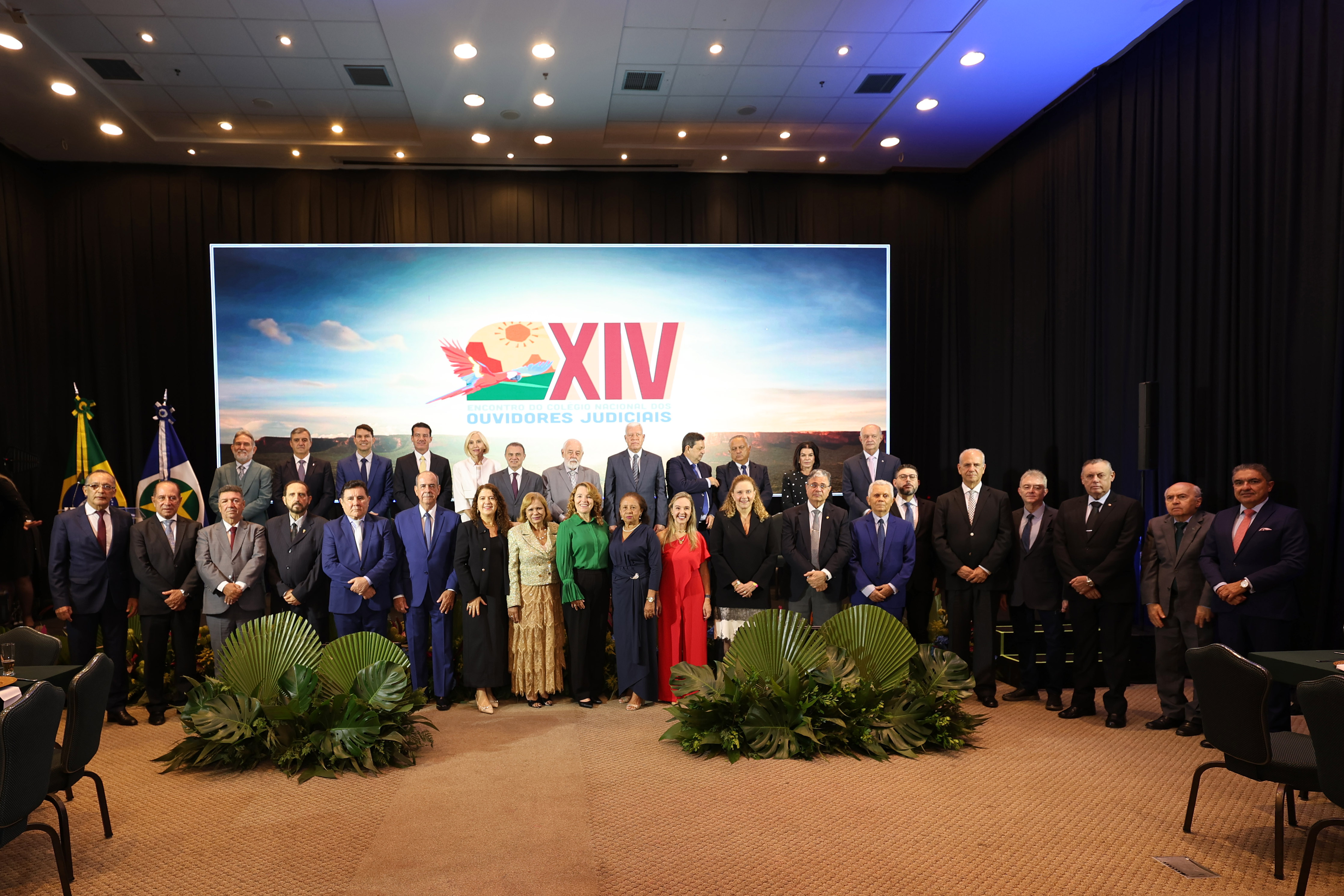 Autoridades posam para foto oficial em palco de evento institucional. Ao fundo, telão exibe a identidade visual do XIV Encontro do Colégio Nacional dos Ouvidores Judiciais. O grupo está organizado em fileiras, com bandeiras ao lado e decoração com plantas na parte frontal do palco.