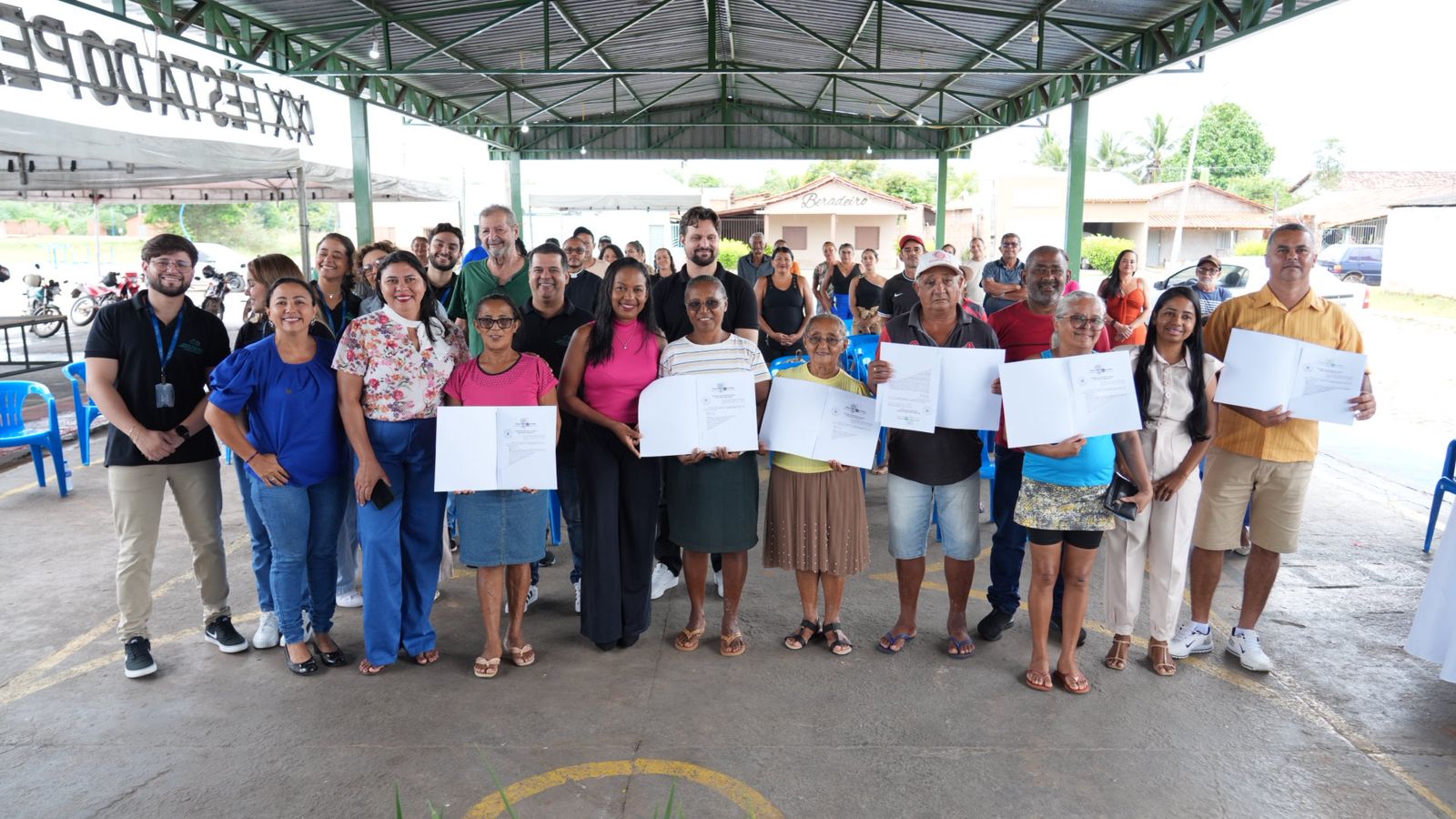 Grupo de pessoas reunidas em um espaço coberto, com estrutura metálica e cadeiras ao fundo, posando para uma foto coletiva. Na frente, homens e mulheres de diferentes idades seguram documentos abertos, aparentando certificados ou registros oficiais. Todos estão sorrindo, transmitindo um clima de celebração e conquista. Ao fundo, outras pessoas acompanham o momento, reforçando o caráter comunitário do evento. O ambiente é simples e aberto, com casas e árvores visíveis ao redor, sugerindo uma ação realizada em uma comunidade local.