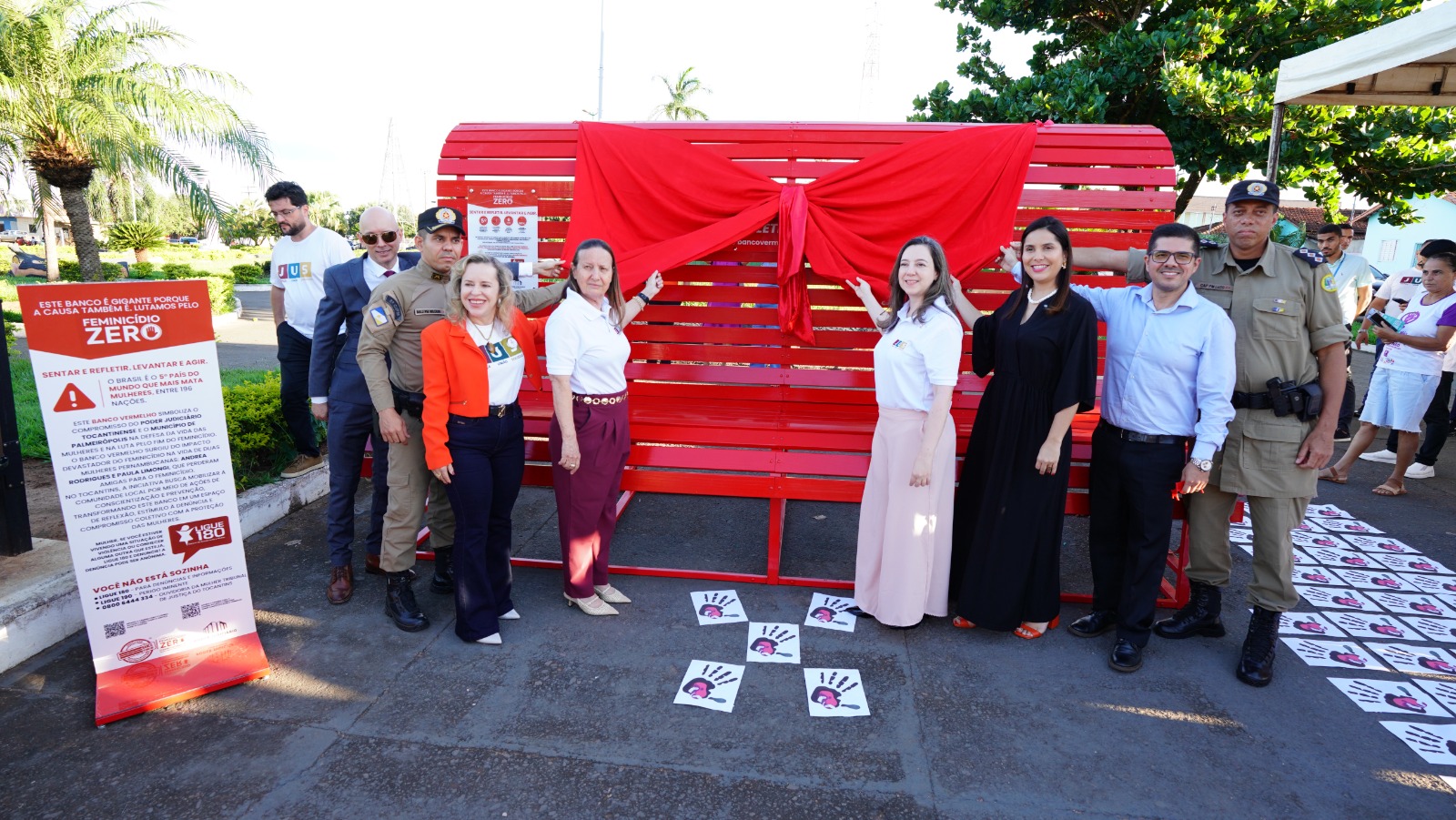 Imagem registra um momento institucional ao ar livre, com um grupo de autoridades e representantes da comunidade reunidos ao redor de um banco vermelho, ainda coberto por um tecido, prestes a ser inaugurado