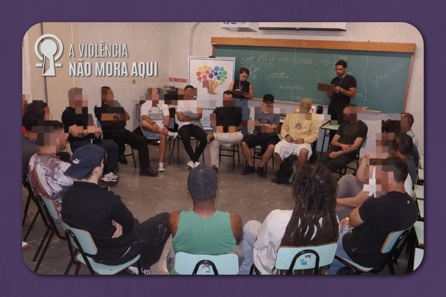 A imagem mostra um grupo de pessoas sentadas em círculo em uma sala, participando de uma roda de conversa mediada por dois facilitadores ao fundo. No ambiente, há um quadro e um cartaz, além da frase “A violência não mora aqui”.