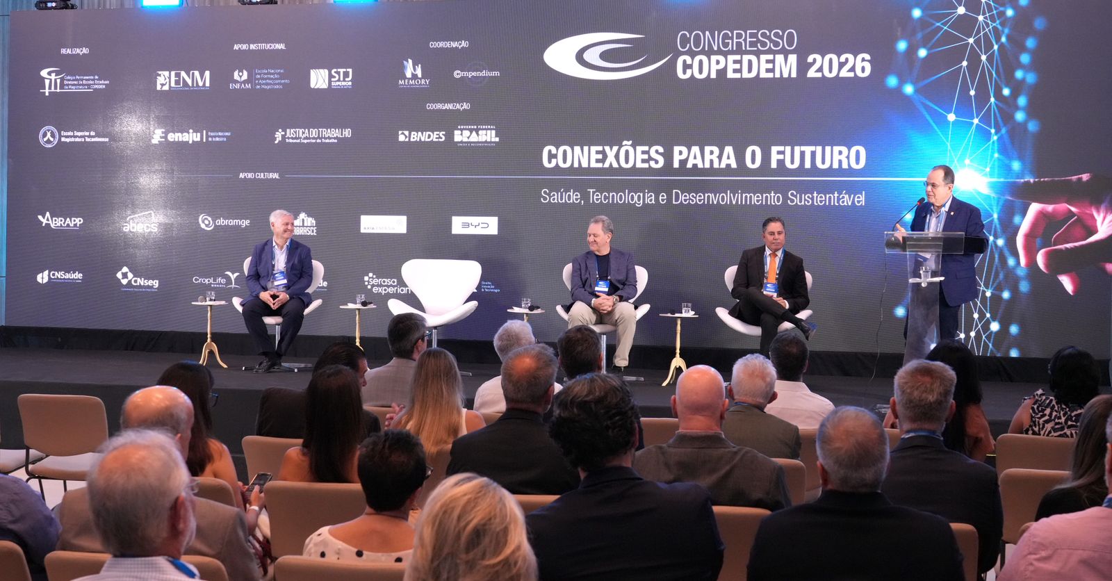 A imagem mostra a abertura do Copedem, com quatro autoridades no palco, sendo uma delas falando ao microfone em um púlpito, enquanto o público acompanha sentado na plateia, de costas. Ao fundo, um grande banner destaca as frases “Congresso Copedem 2026”, “Conexão para o futuro” e “Saúde, tecnologia e desenvolvimento sustentável”.