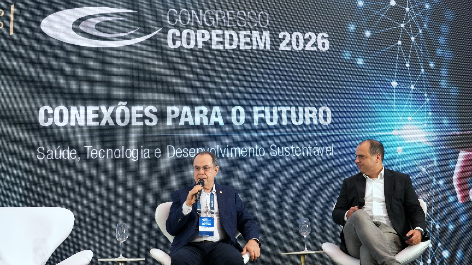 A imagem mostra dois homens sentados em poltronas brancas durante um painel do Congresso COPEDEM 2026, em um palco com telão ao fundo. Um deles fala ao microfone enquanto o outro observa, com a temática “Conexões para o Futuro” destacada atrás.