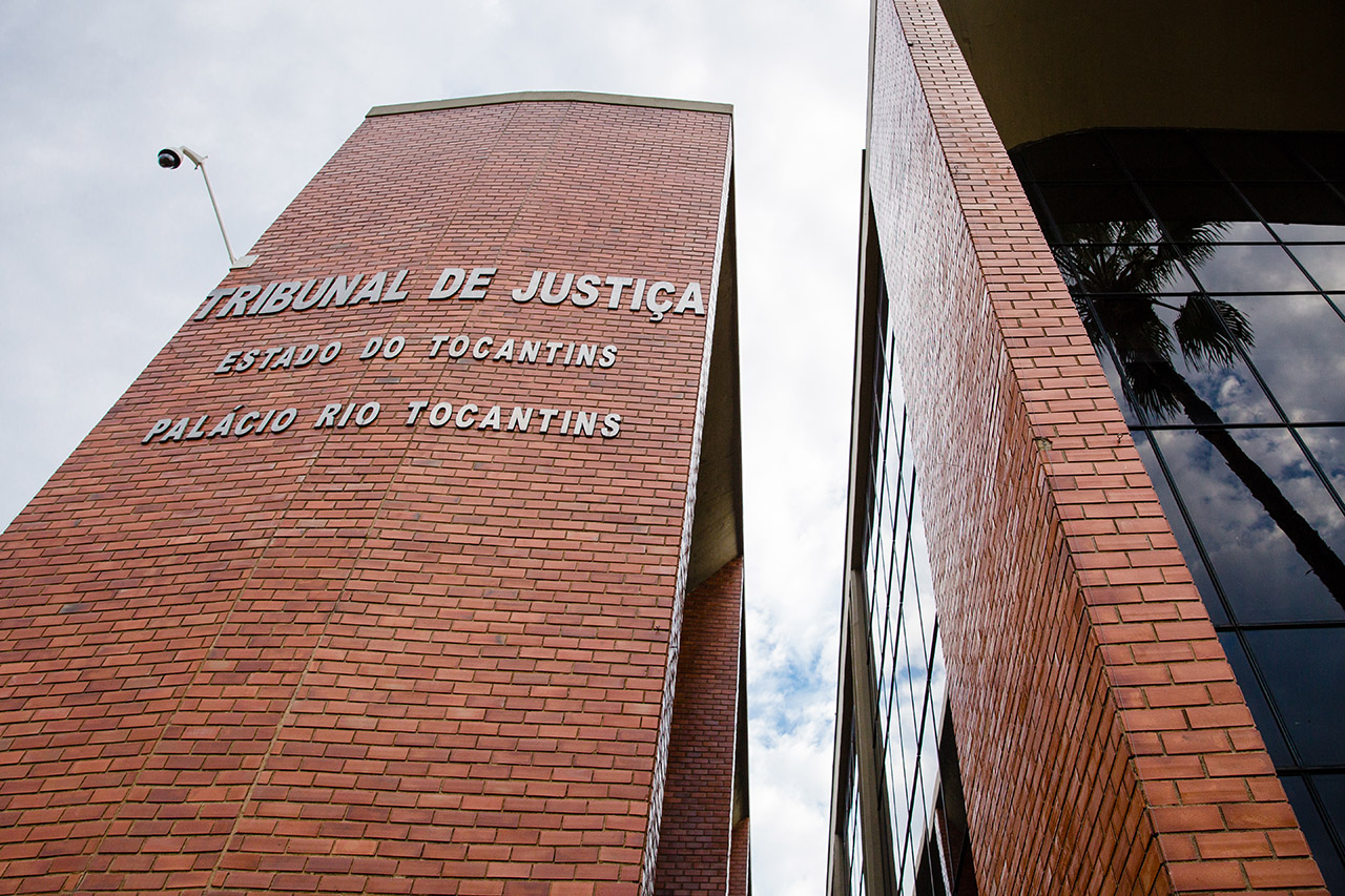 Fachada do Tribunal de Justiça do Tocantins, com revestimento em tijolos aparentes e inscrições em letras metálicas. À direita, estrutura com vidro espelhado refletindo o céu e uma palmeira. No topo, há uma câmera de segurança.