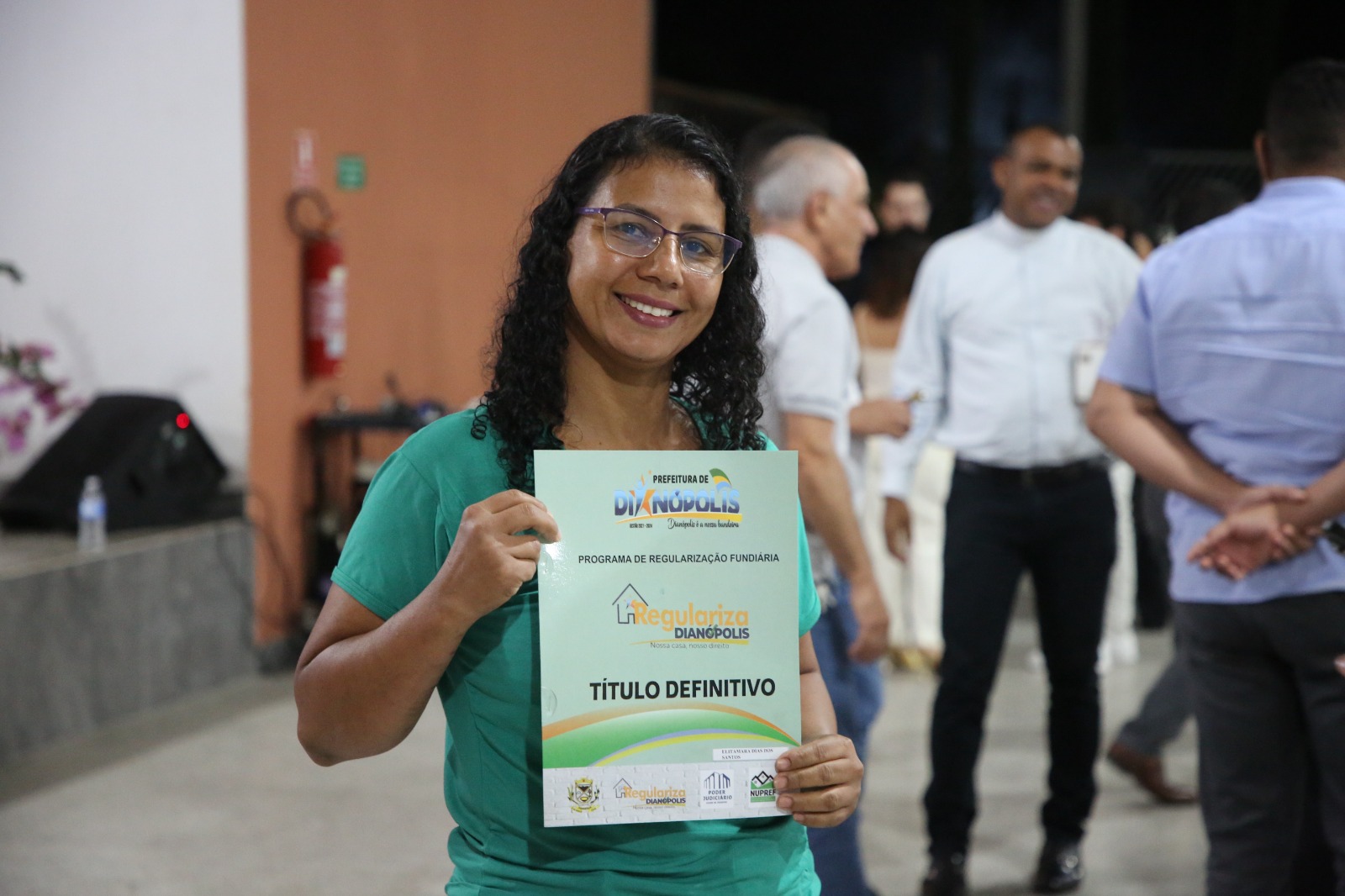 A imagem mostra uma mulher sorrindo enquanto segura um documento de “Título Definitivo” do programa de regularização fundiária de Dianópolis. Ao fundo, outras pessoas conversam em um ambiente interno.